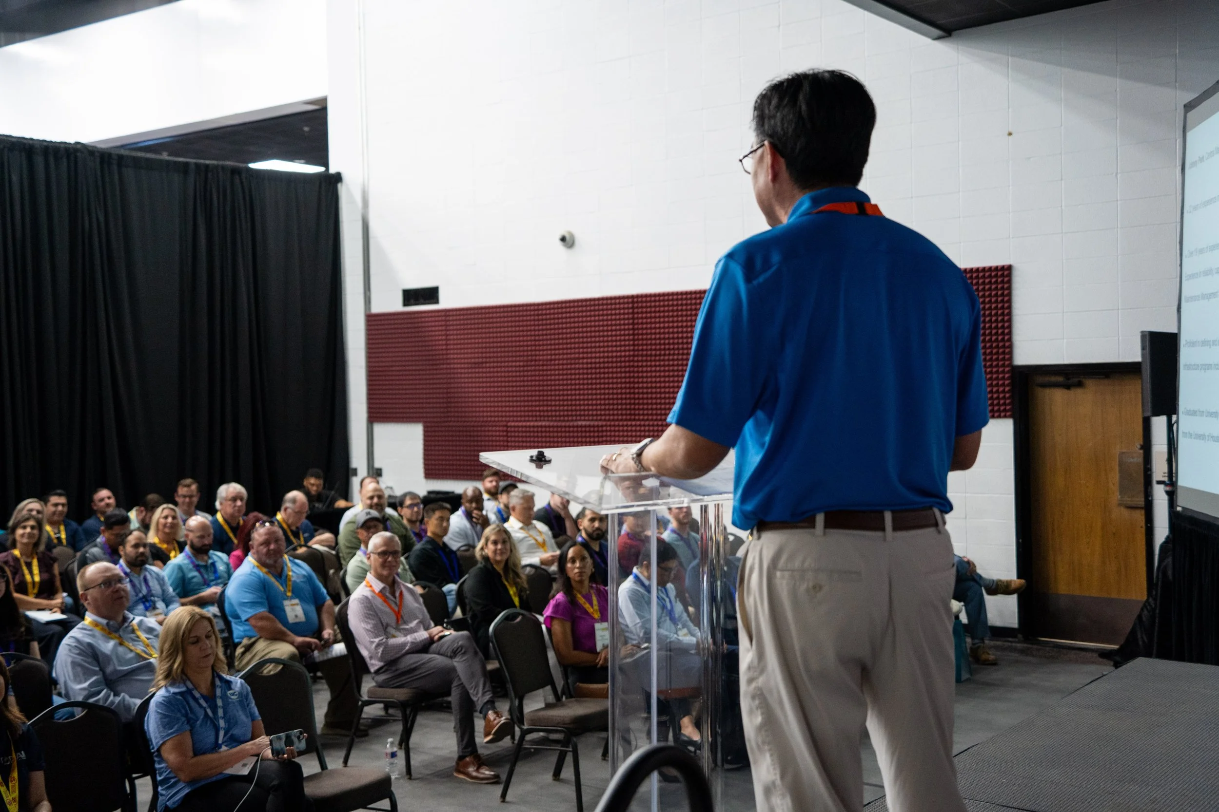 A man in a blue shirt giving a presentation to an audience in a conference room.