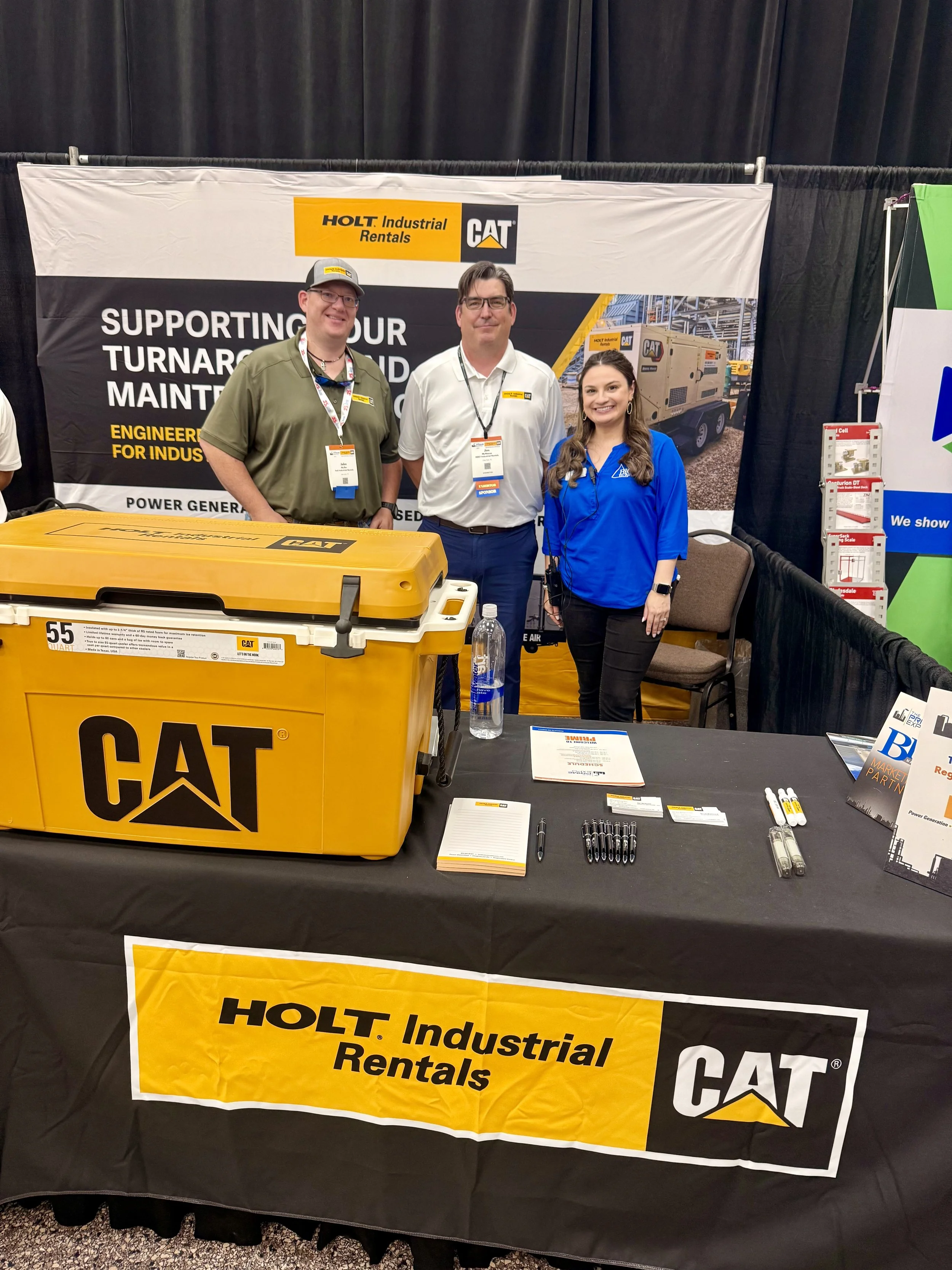 Three people standing behind a booth at a trade show for Holt Industrial Rentals, a Caterpillar company. The booth has promotional materials, a yellow CAT container, and a banner with the company's logo.