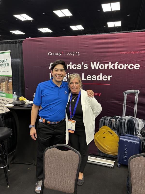 Two people, a man and a woman, standing together at a conference booth with a maroon backdrop that reads 'America's Workforce' and 'Corpay Lodging.' The man is wearing a blue polo shirt and black pants, and the woman is wearing a white blazer over a 