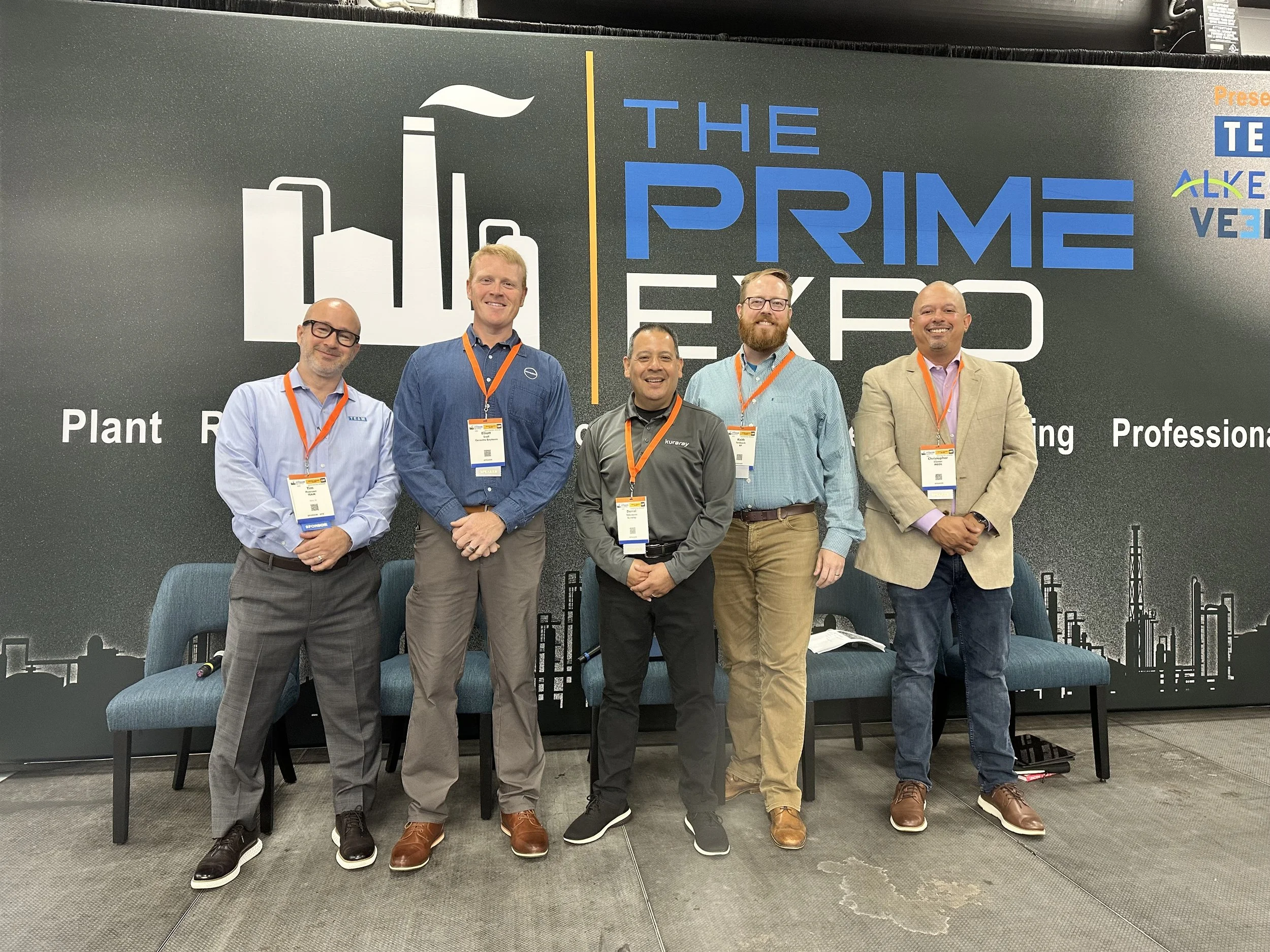 Five men are standing in front of a large display at The Prime Expo. They are smiling and wearing conference badges with orange lanyards. The background features a graphic of factory buildings and text related to the expo.