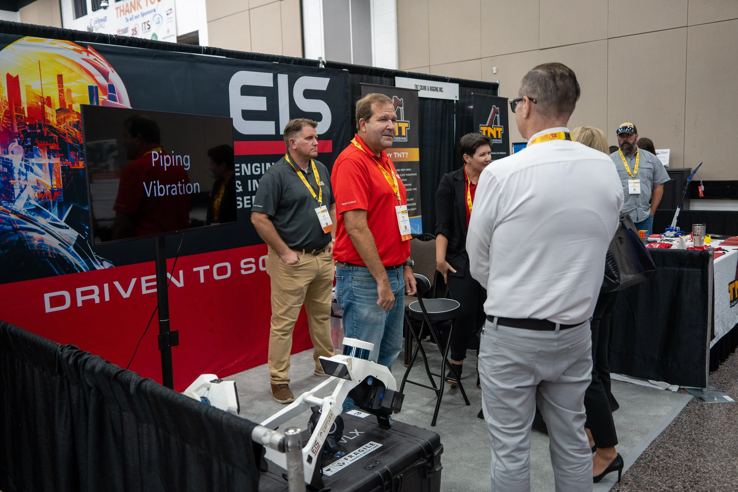 People attending an exhibition booth with a large display screen showing 'Piping Vibration' and a cylindrical robotic arm in front of it. Several individuals are engaged in conversation or viewing exhibits.