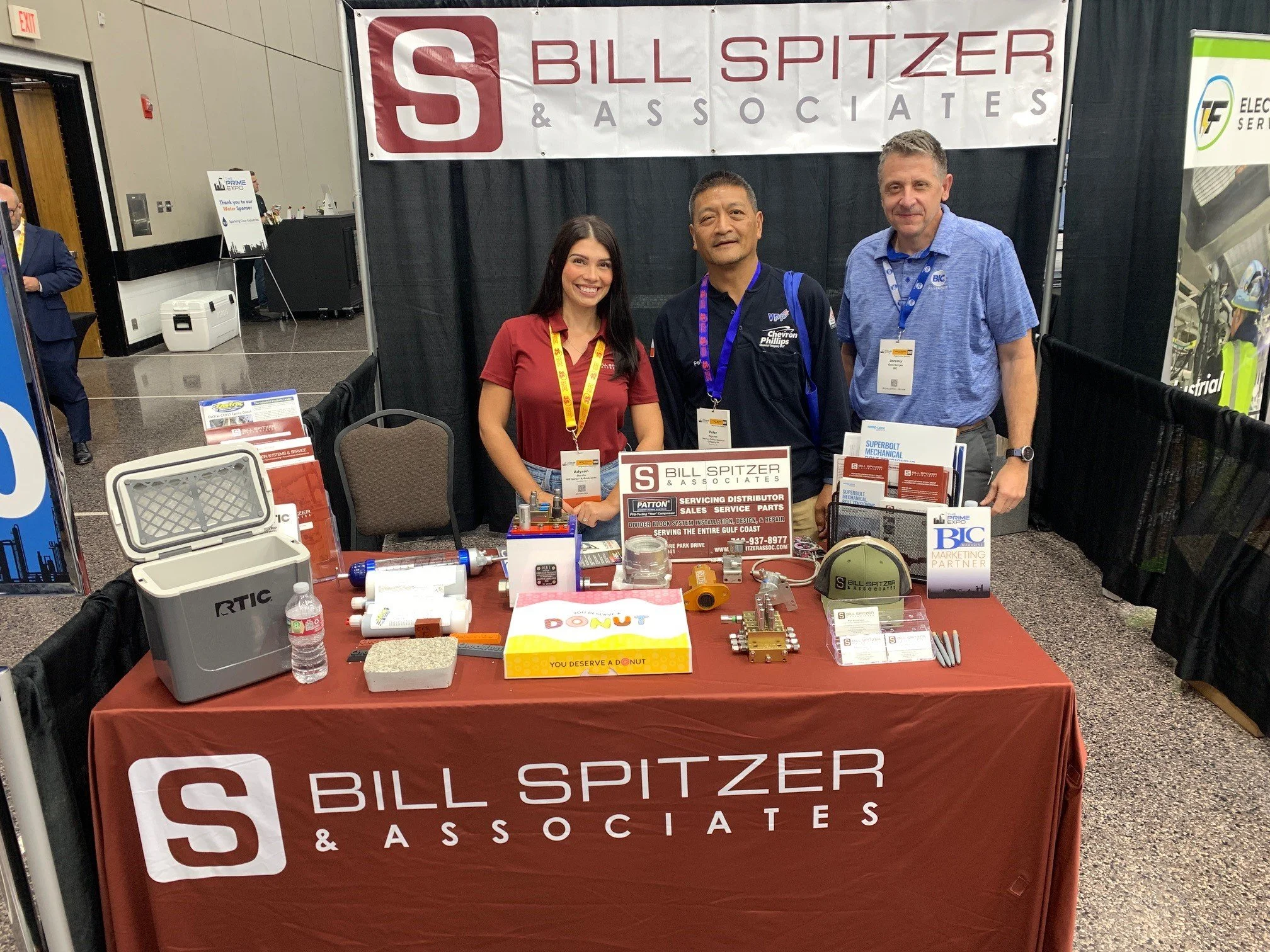 Three people standing behind a booth with a red tablecloth that has the logo and name 'Bill Spitzer & Associates'. The booth displays various mechanical parts and promotional materials. The background includes a large banner with the same logo and na