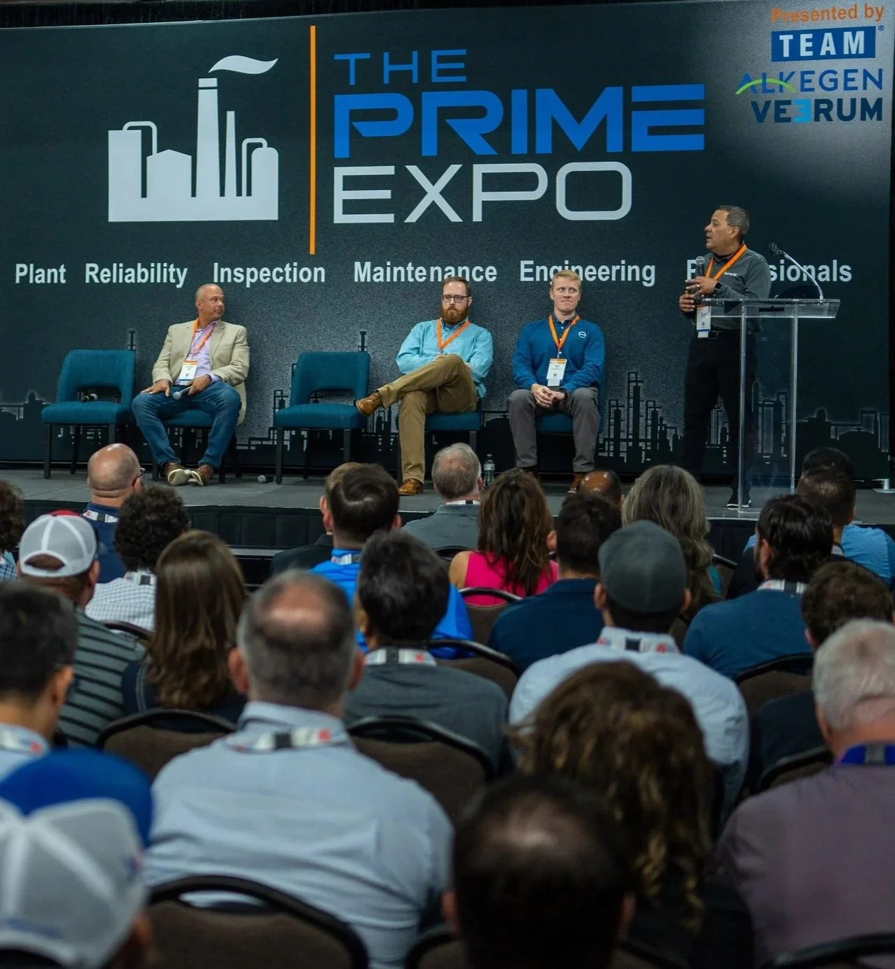 Panel discussion at The Prime Expo with four men on stage, one speaking at a podium, and an audience watching, with a large screen behind displaying the event's name and topics like plant reliability, inspection, maintenance, engineering, and professionals, presented by TEAM ALGEN VEIRUM.