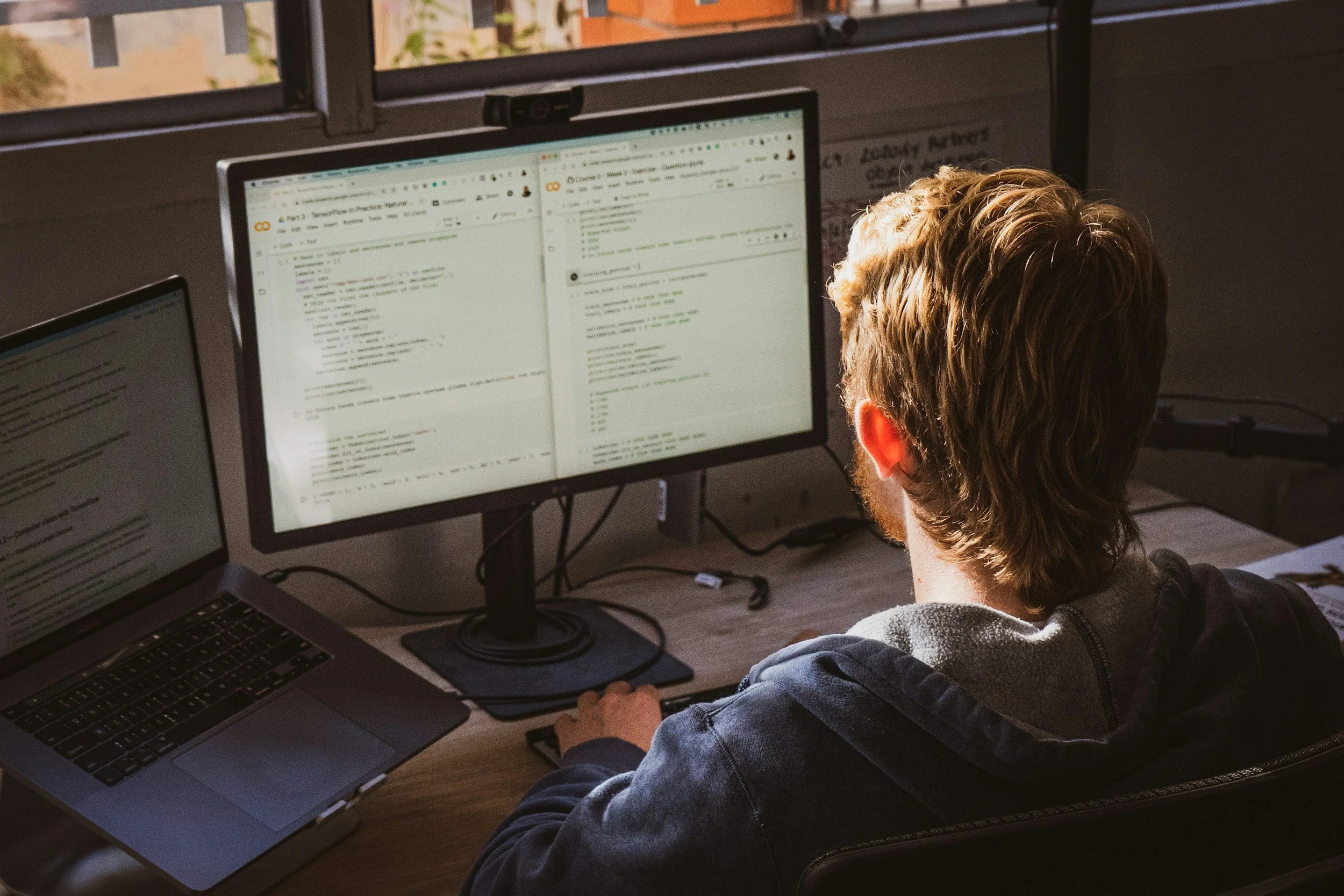 A person with short brown hair working at a desk with multiple monitors, typing on a keyboard, and viewing lines of code and text on the screens in a room with open windows.