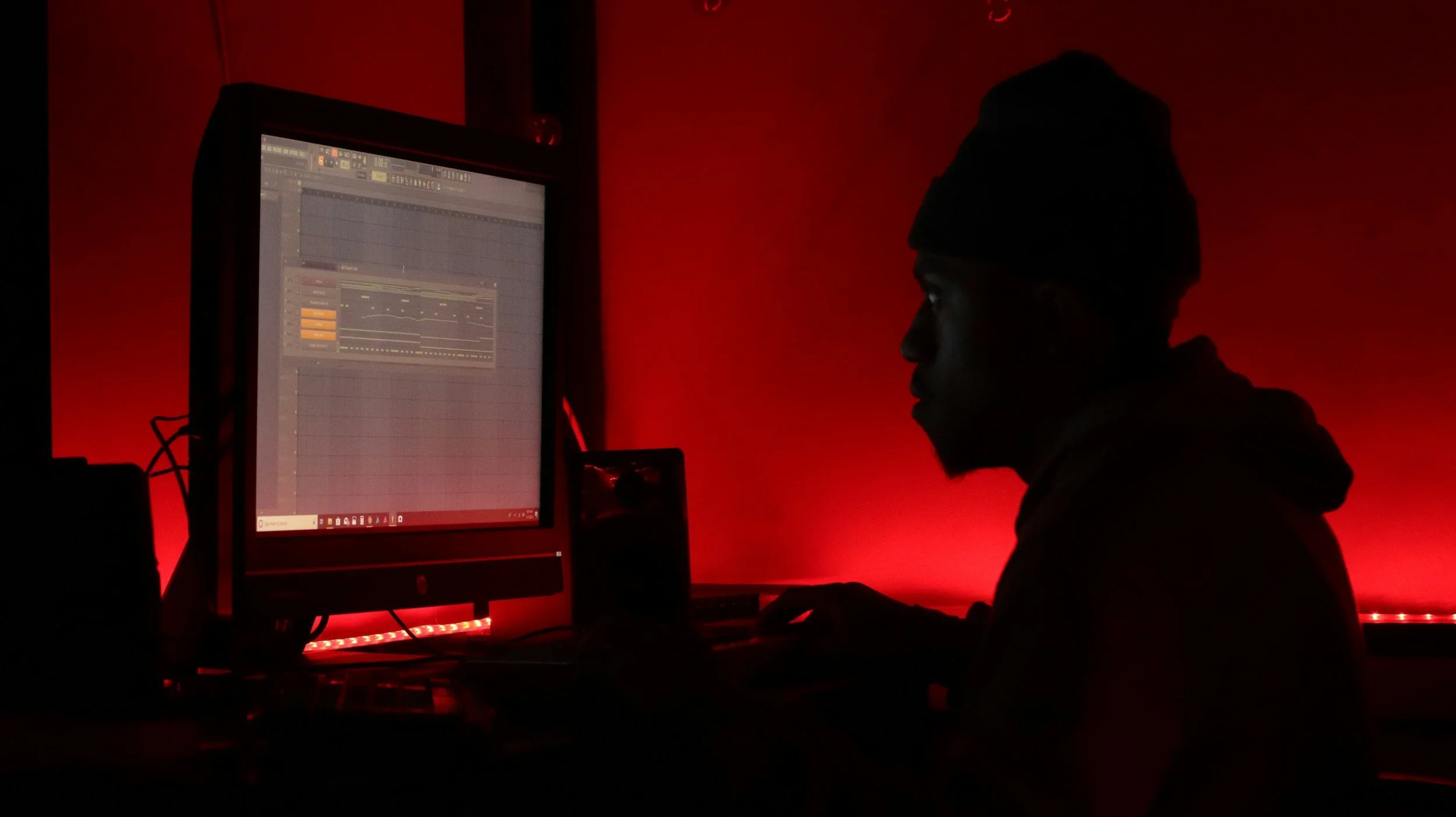 Person wearing a beanie working on a music production computer in a dark room with red lighting.