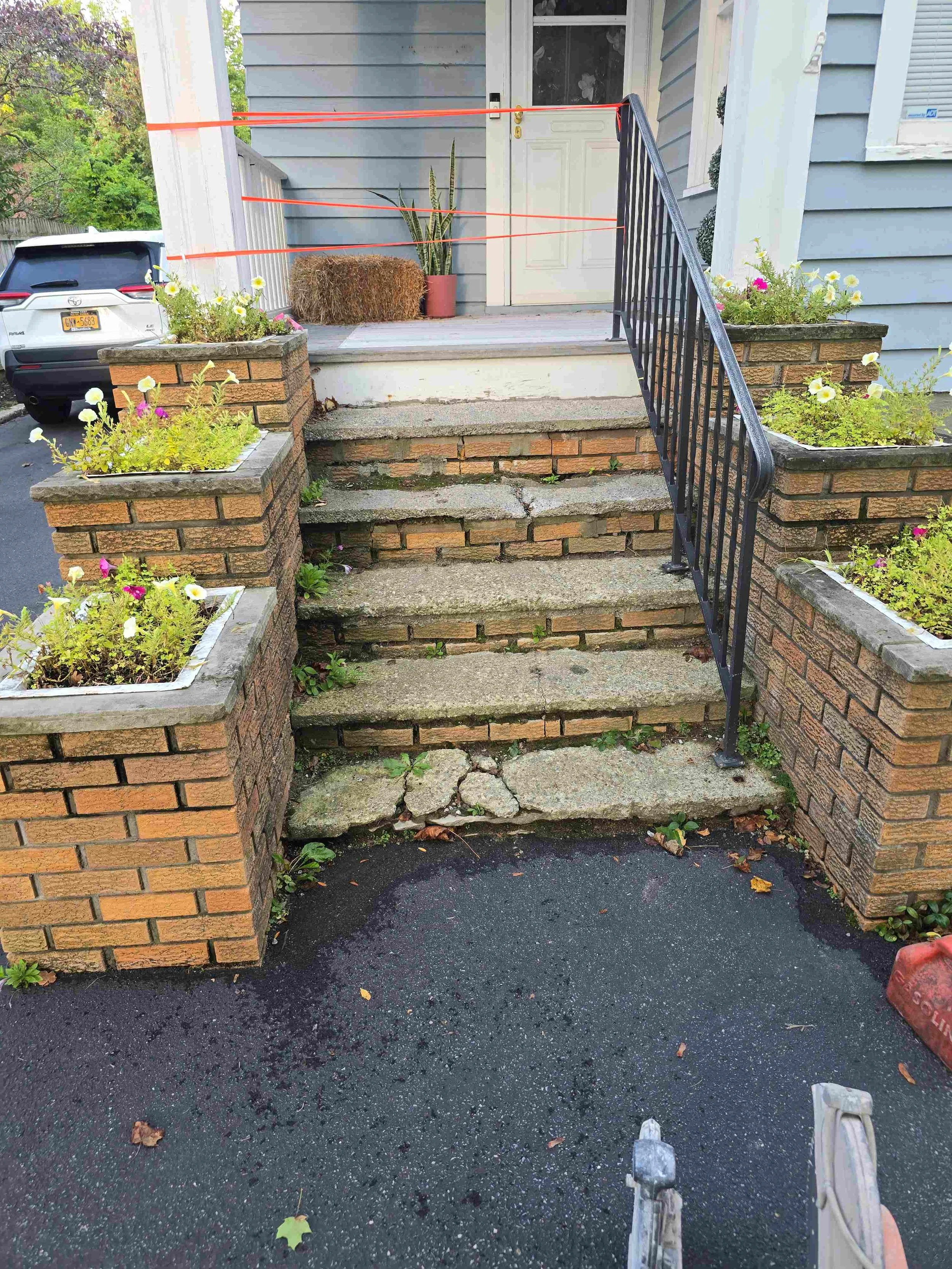 A front porch with a set of steps leading up to a door. The steps are cracked and uneven. The porch is decorated with potted plants and flowers in planters on the brick sides of the staircase. An orange plastic barrier is stretched across the doorway