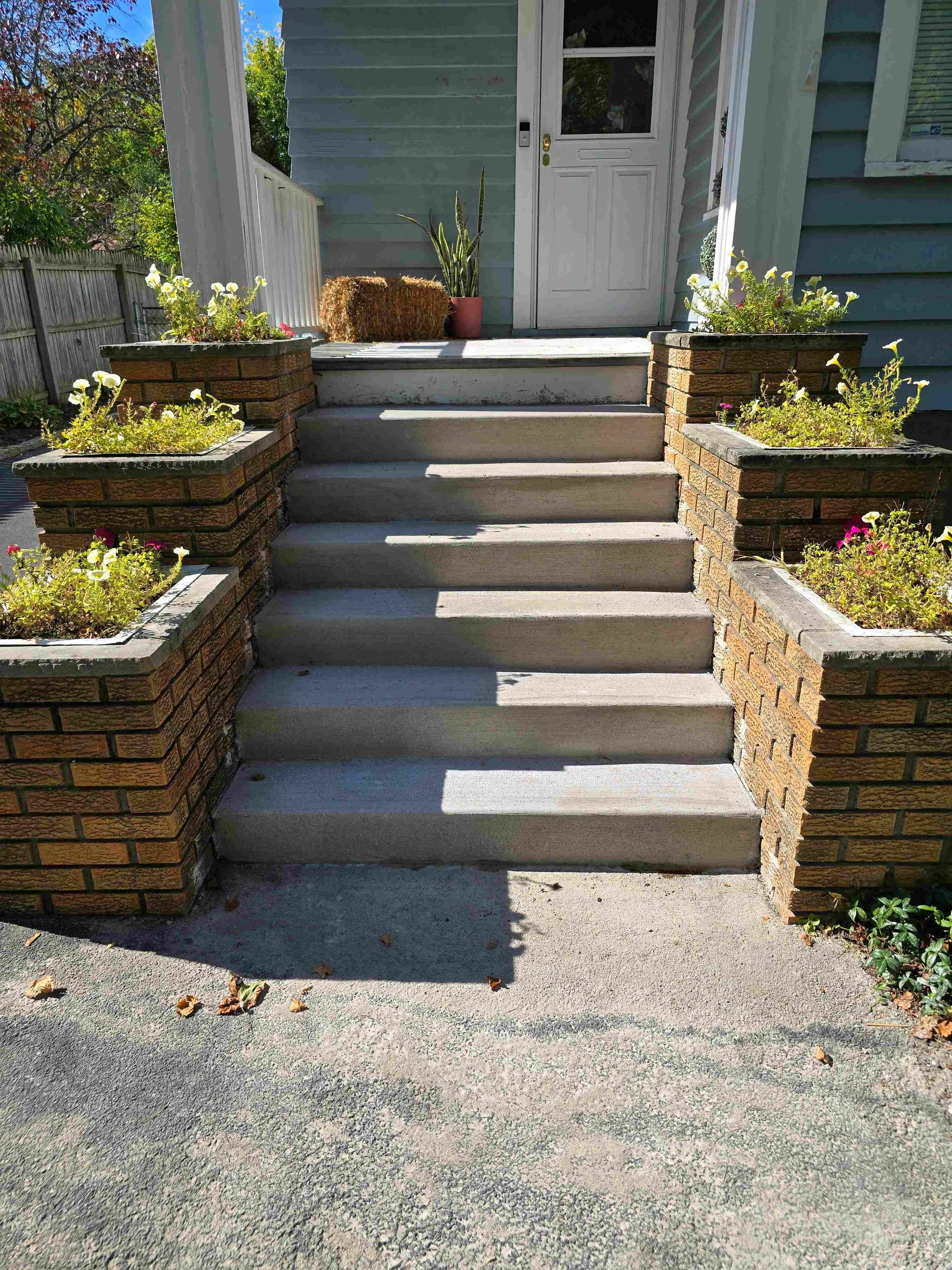 Front steps leading to a house entrance with flower planters on both sides of the steps, a white door, and a doormat. The area is sunny with a clear blue sky, trees, and a wooden fence in the background.