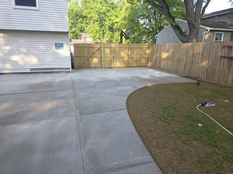 Empty backyard with concrete patio, grass area, wooden fence, house wall, and a basketball hoop on the fence.
