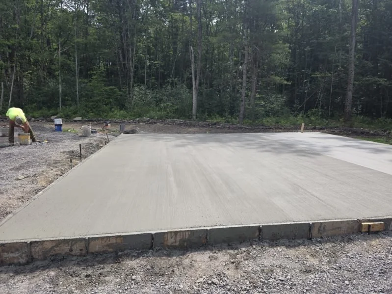 Freshly poured concrete slab on a construction site surrounded by trees, with a worker on the left smoothing the surface.
