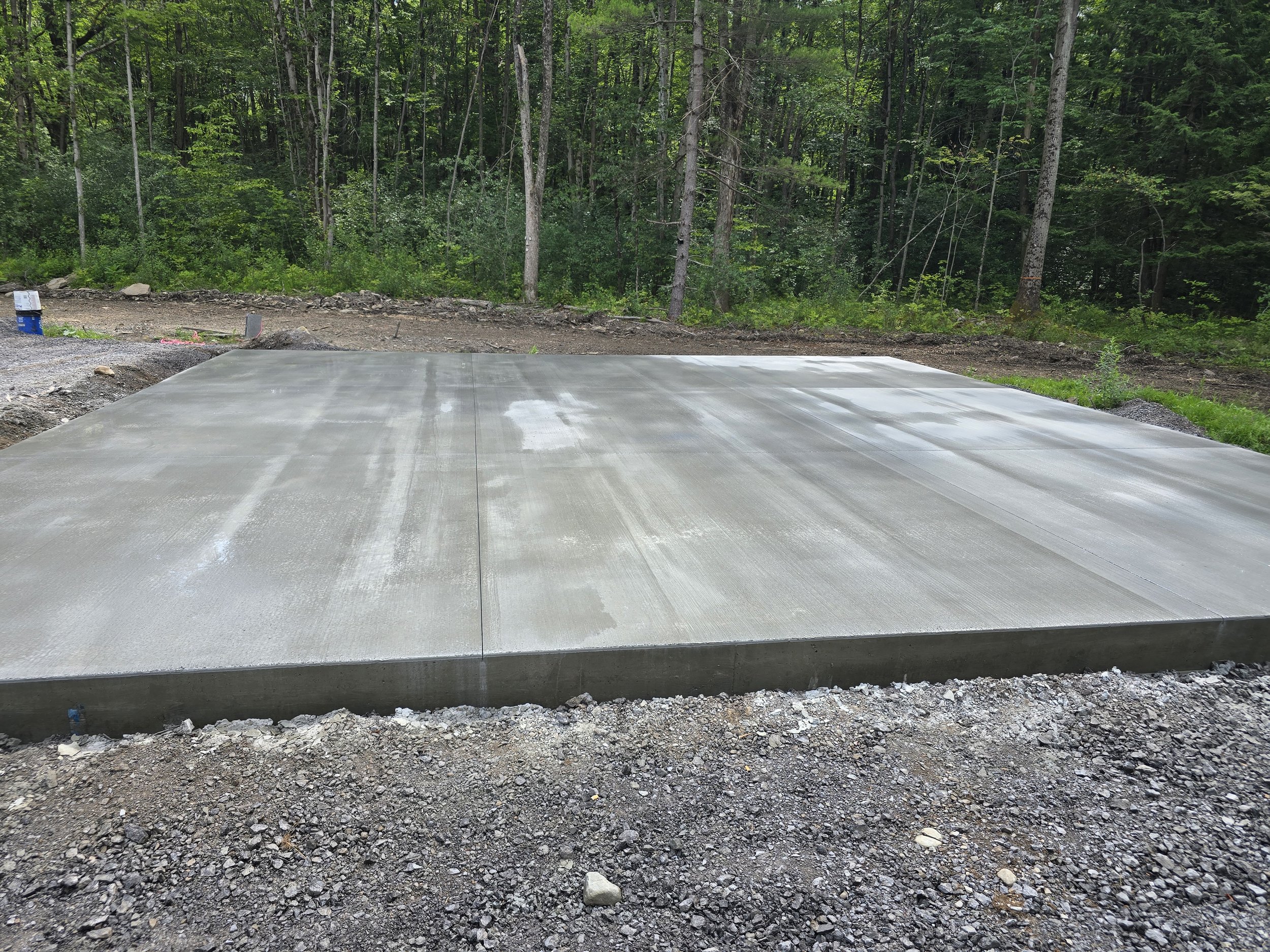 Freshly poured concrete slab in a wooded area, with a dirt road and trees in the background.