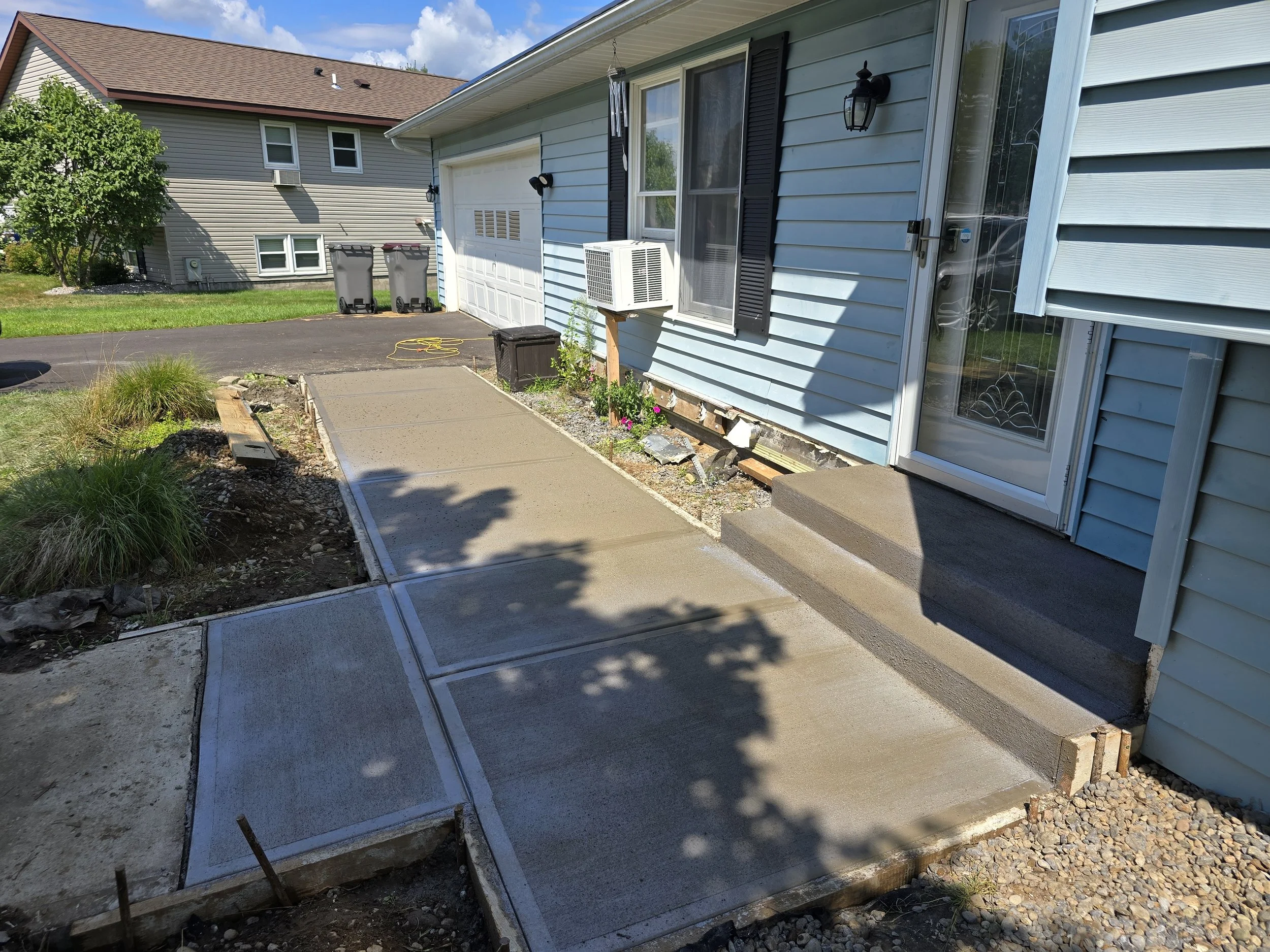 Newly poured concrete sidewalk in a residential backyard with a house, patio, and two trash bins in the background.