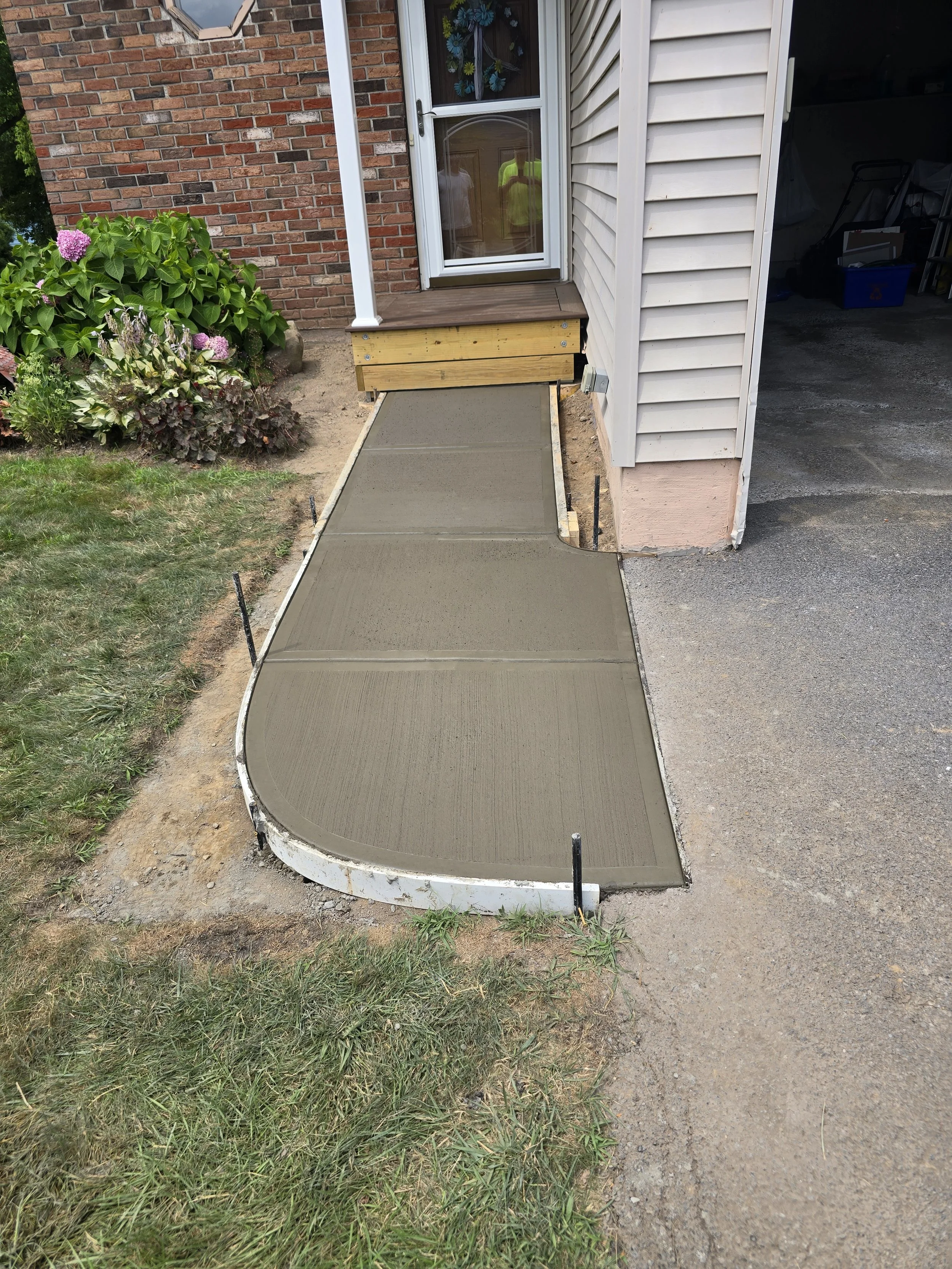 New concrete sidewalk in front of house with brick and vinyl siding exterior, garden with pink hydrangea plants, and a partially open garage with storage items.