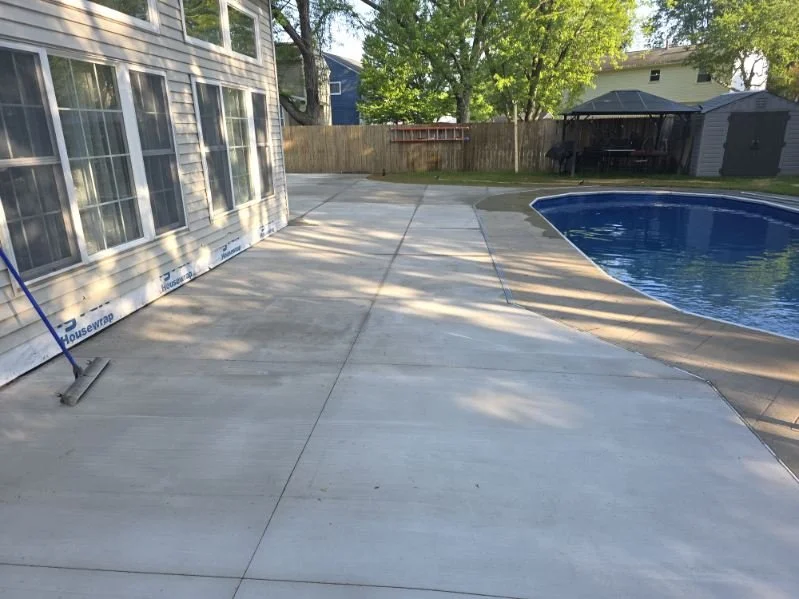 Backyard with concrete patio, swimming pool with blue water, wooden fence, shed, and trees in the background