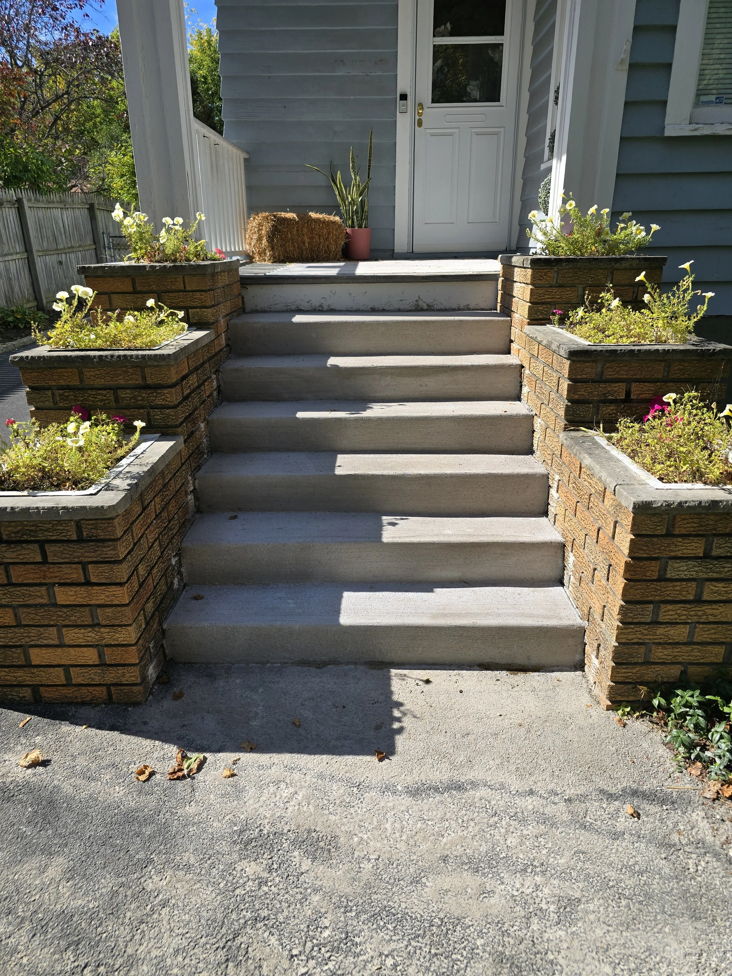 Concrete steps leading up to a house front door, flanked by brick planters with potted plants and flowers, with a wooden fence and trees in the background.