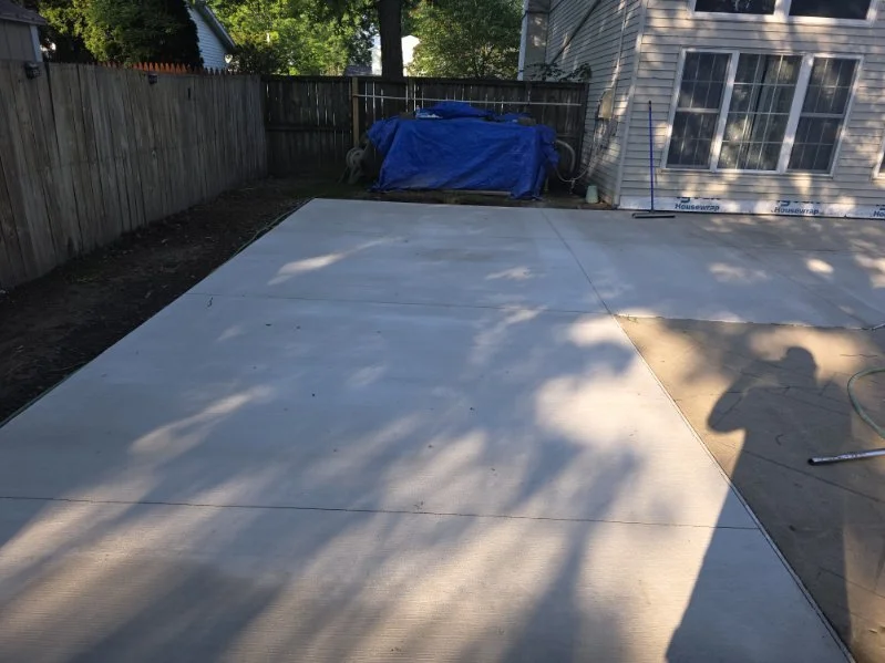 A backyard patio under construction with a concrete slab, a wooden fence, a house with siding, and construction tools and materials, including a blue tarp covering items in the background.