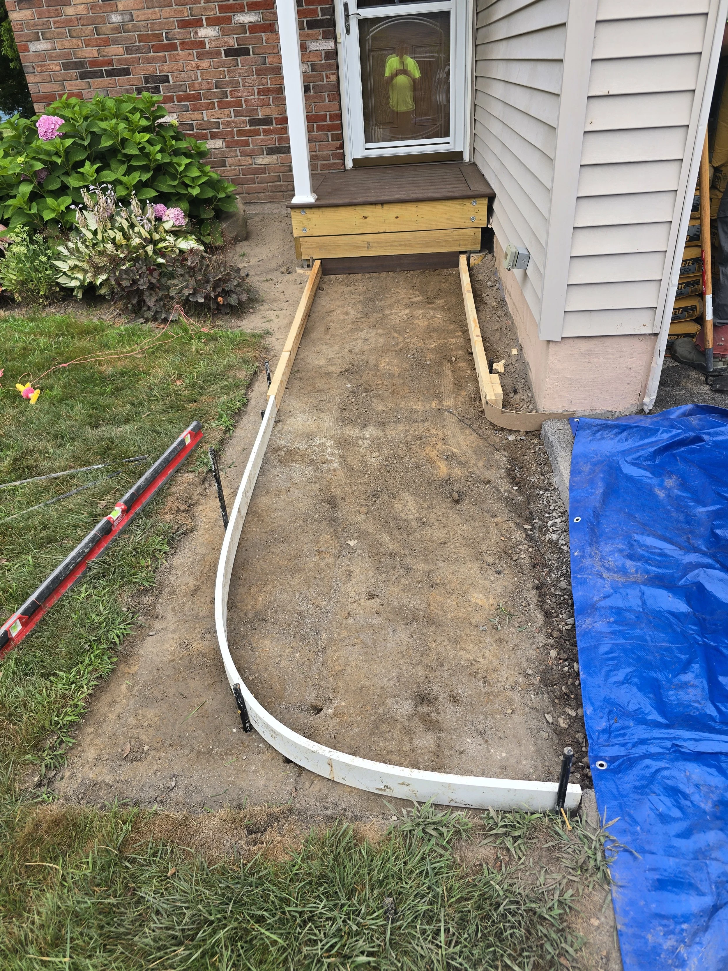 A backyard with a flower garden on the left and a house entrance on the right, where a small wooden platform with a dot in the center is being installed, with tools and blue tarp on the ground.