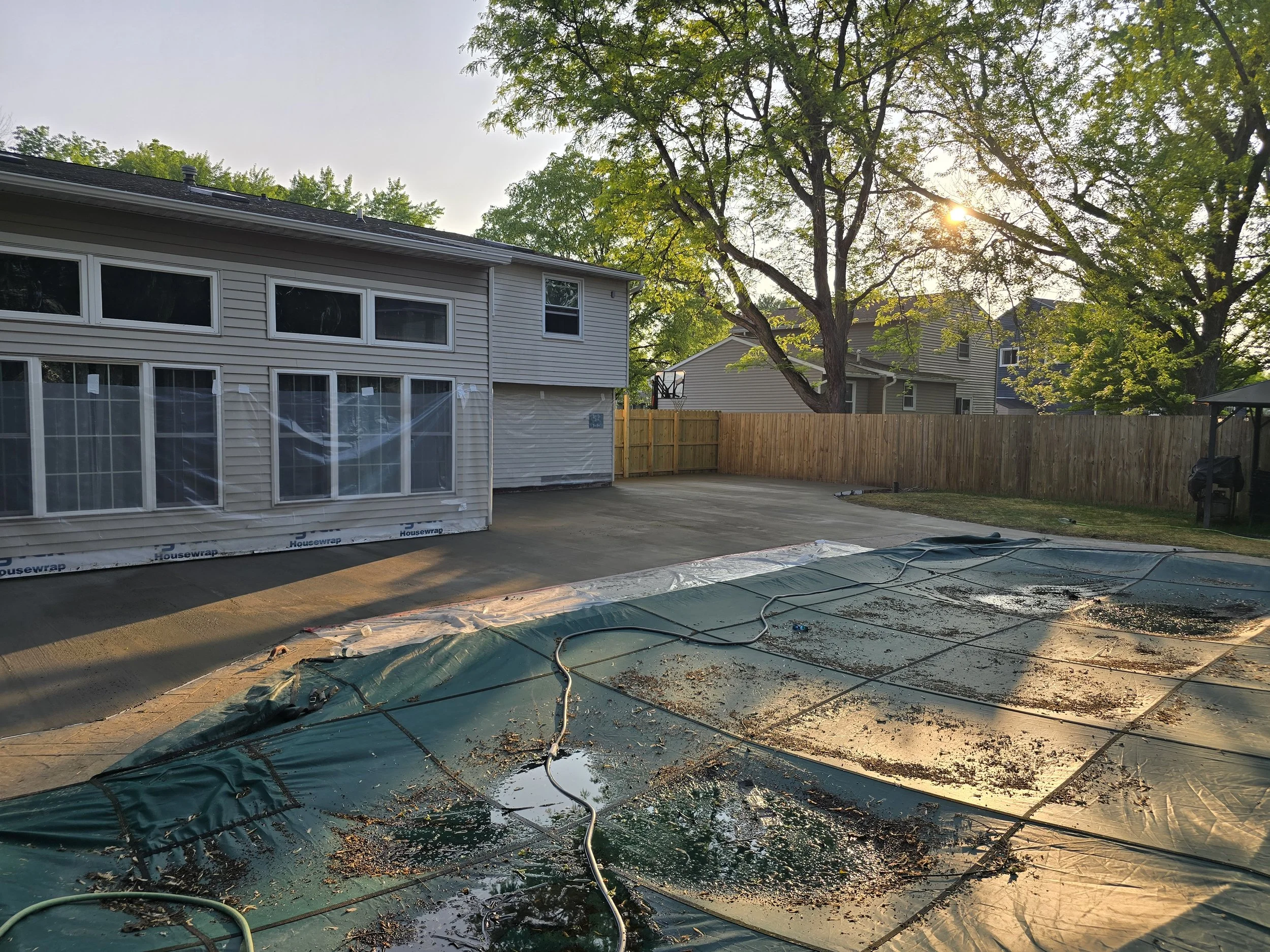 A backyard scene during sunset with a house under construction or renovation, with scaffolding, plastic sheeting on the ground, a large tree with green leaves, and neighboring houses visible in the background.