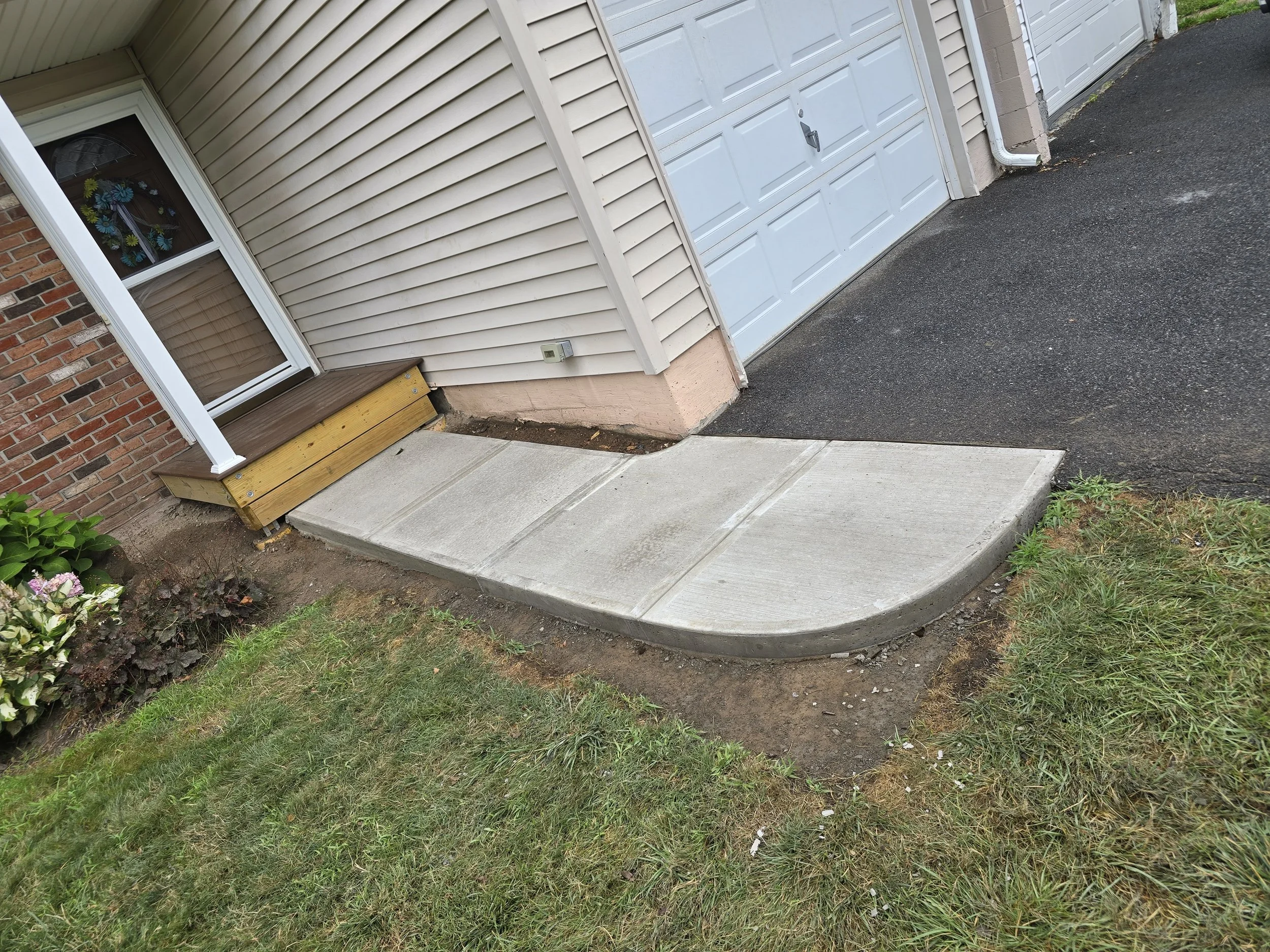 A suburban house entrance with a small concrete patio, a yellow wooden step, a flower decoration on the front door window, and a white garage door on the side.