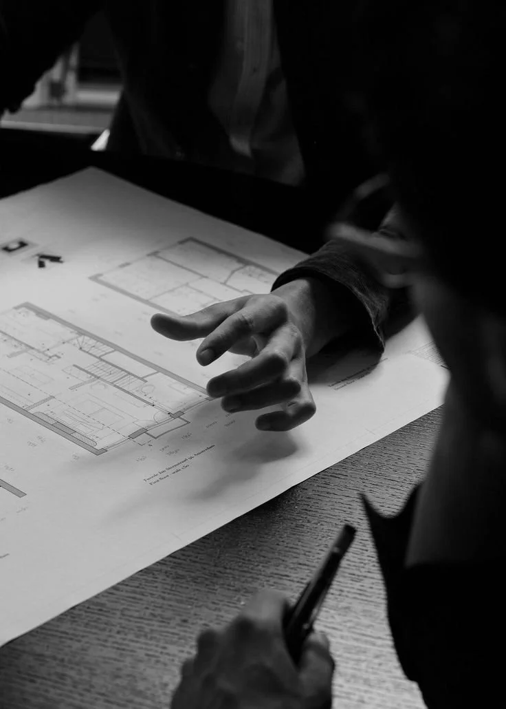 A person pointing at a printed architectural floor plan on a table, with another person holding a pen nearby.