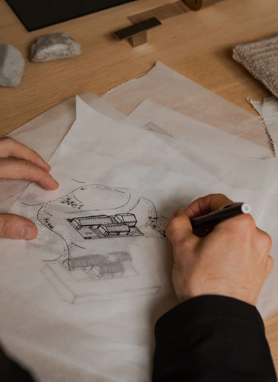 Person sketching a house or building design on tracing paper with a pen, with architectural drawings and fabric samples on a wooden table.