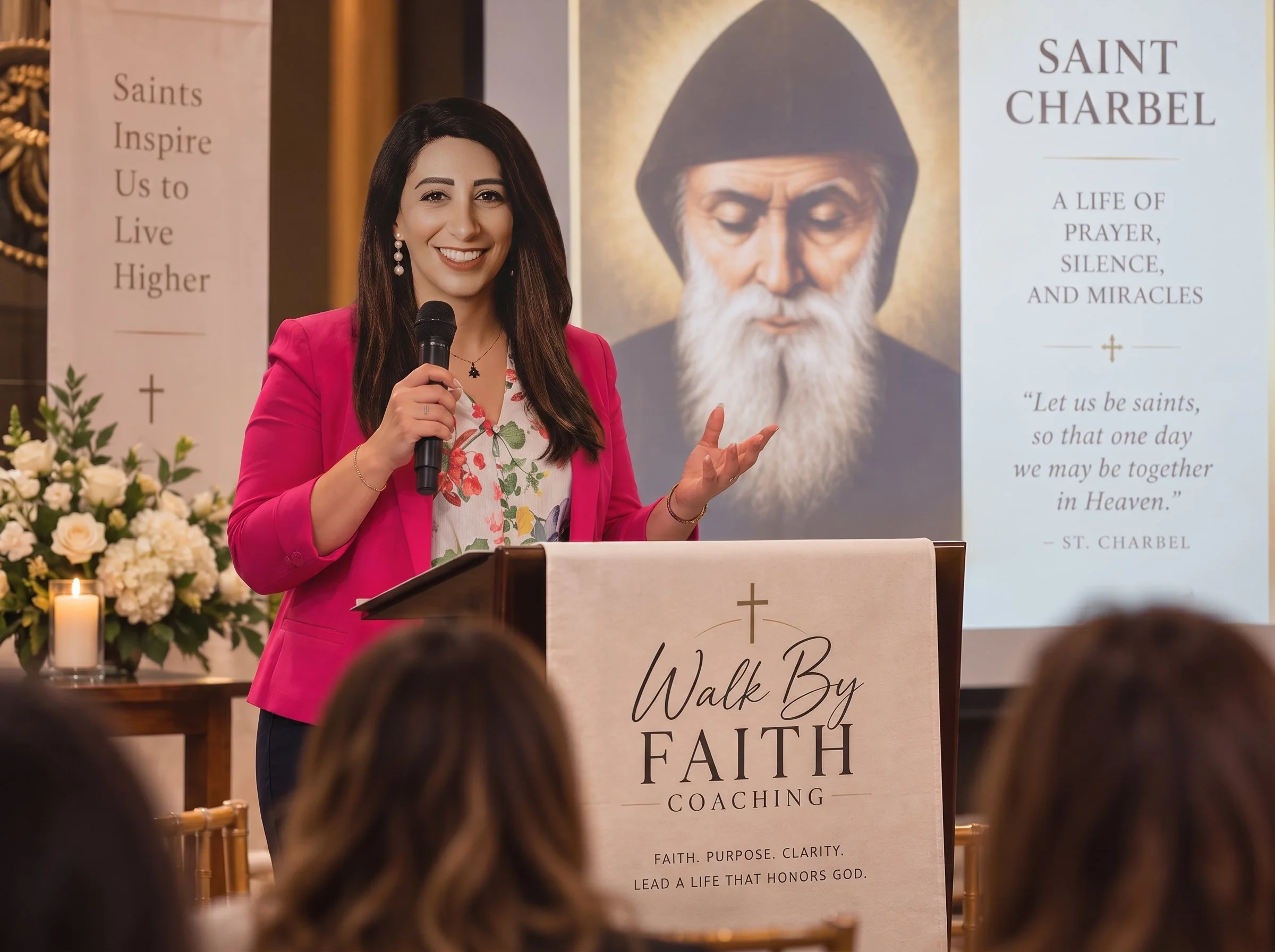 A woman in a pink blazer and floral blouse is speaking into a microphone during a church event. Behind her there is a large banner with a portrait of Saint Charbel and text about prayer, silence, and miracles. A floral arrangement and a lit candle are visible in the foreground.