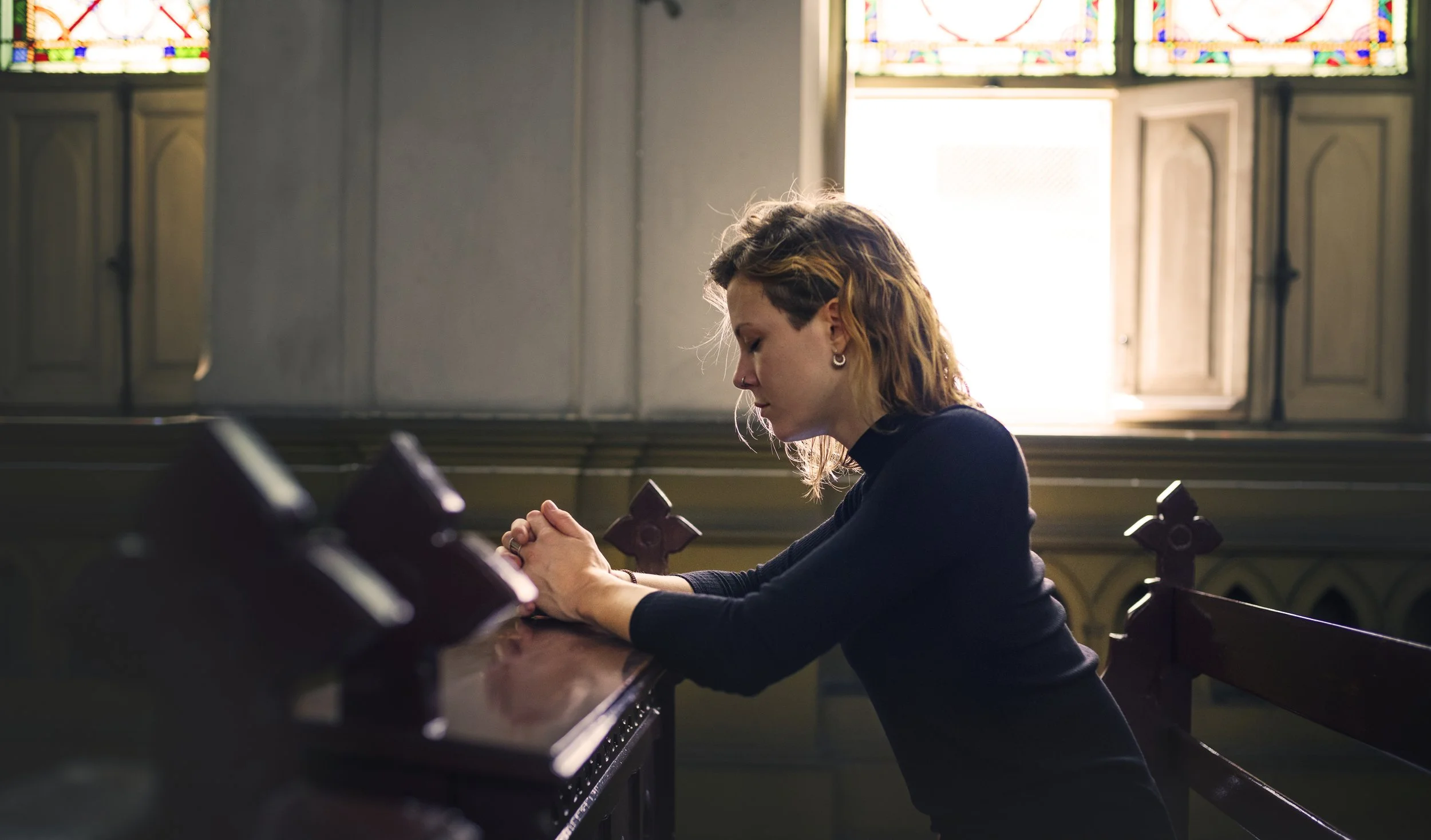 A woman sitting at a church pew with her hands clasped in prayer, head bowed, inside a church with stained glass windows in the background.