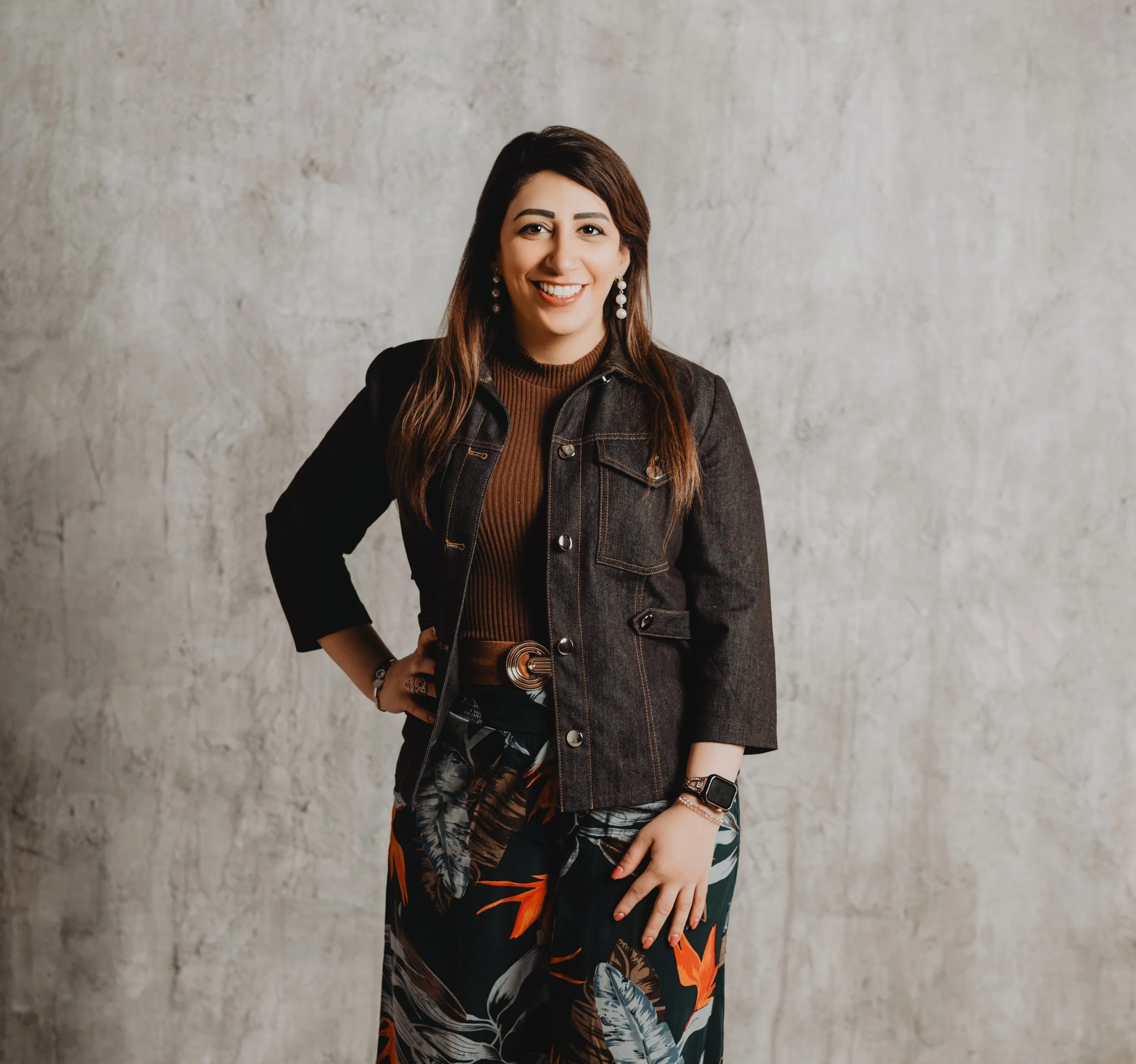 A woman with long brown hair, wearing earrings, a brown top, and a dark denim jacket, stands against a plain gray wall, smiling at the camera.