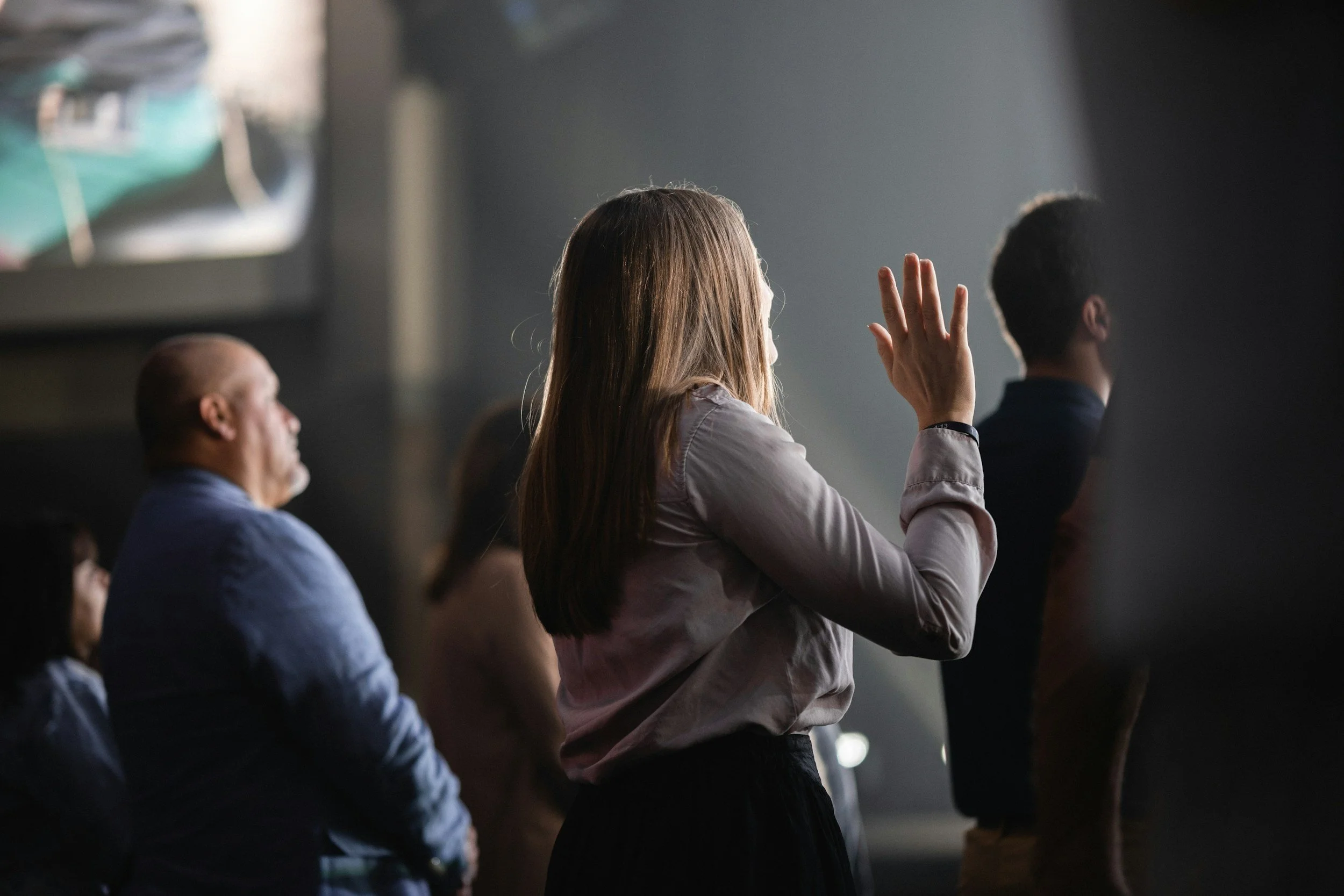 A woman with long hair raising her hand during a meeting or presentation, seated among other people in a dimly lit room.