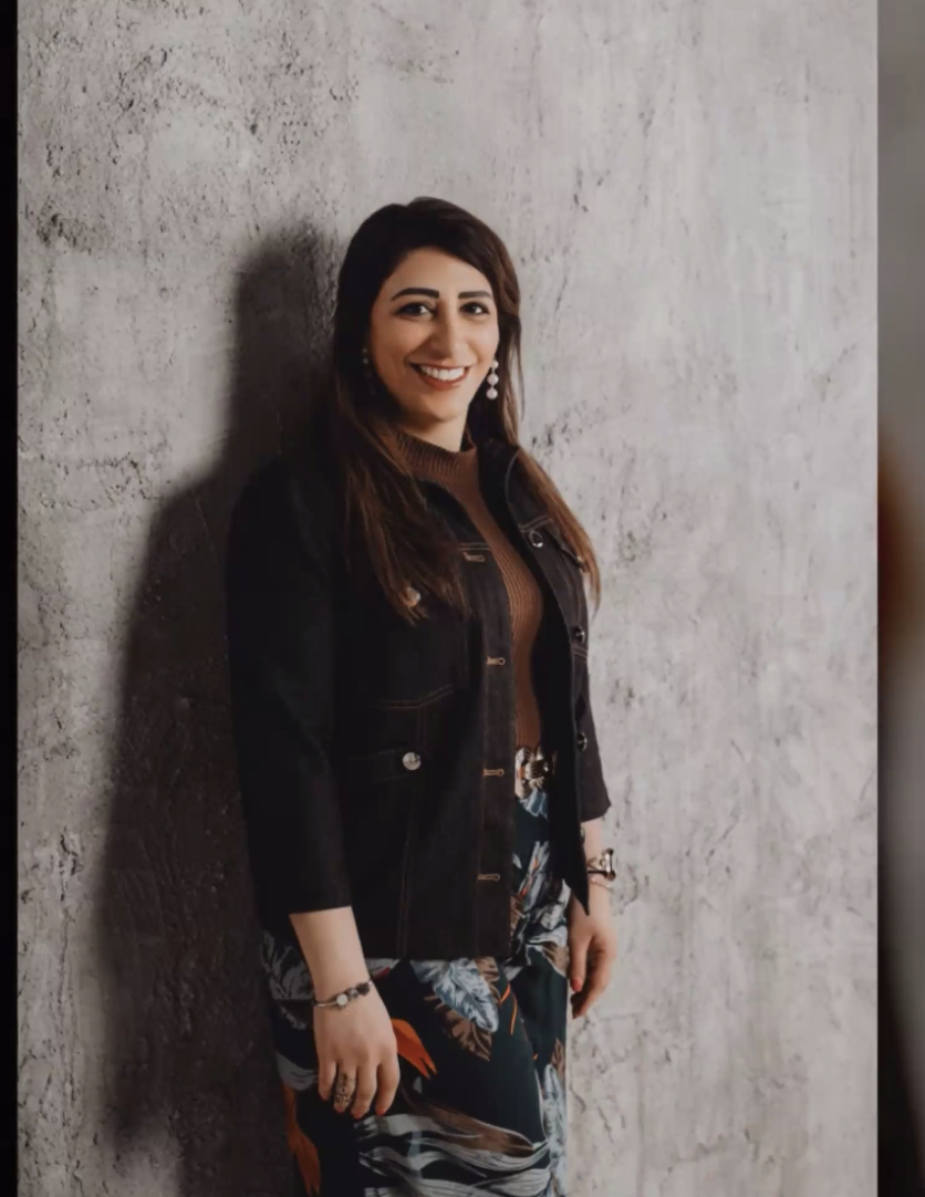 A cheerful woman with long brown hair and earrings, standing against a textured concrete wall, wearing a black jacket over a brown top and a patterned skirt.