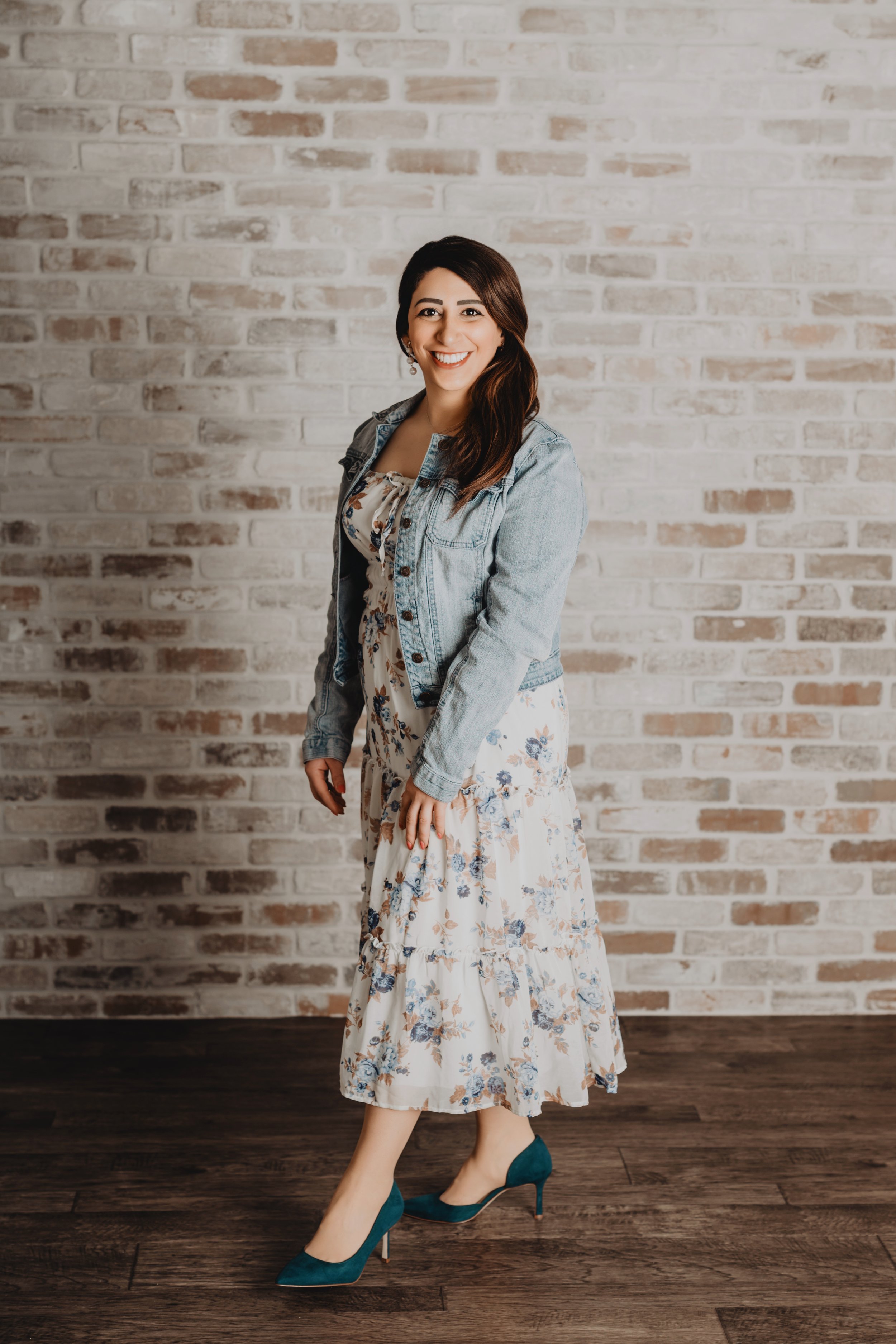 Young woman with dark brown hair smiling, wearing teal high heels, a floral dress, and denim jacket, standing in front of a white brick wall, with dark hardwood floor.