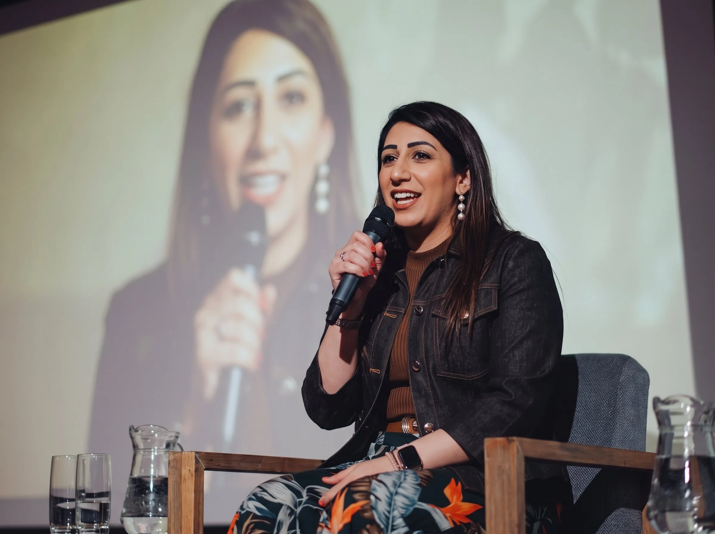 A woman with dark hair, earrings, and a leather jacket speaking into a microphone during a presentation or discussion. She is sitting at a table with glasses of water, with a large projected image of her on a screen behind her.