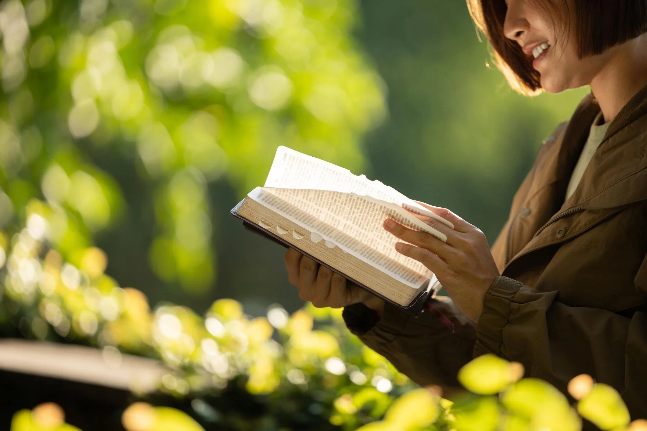 Person reading a book outdoors with trees and sunlight in the background.