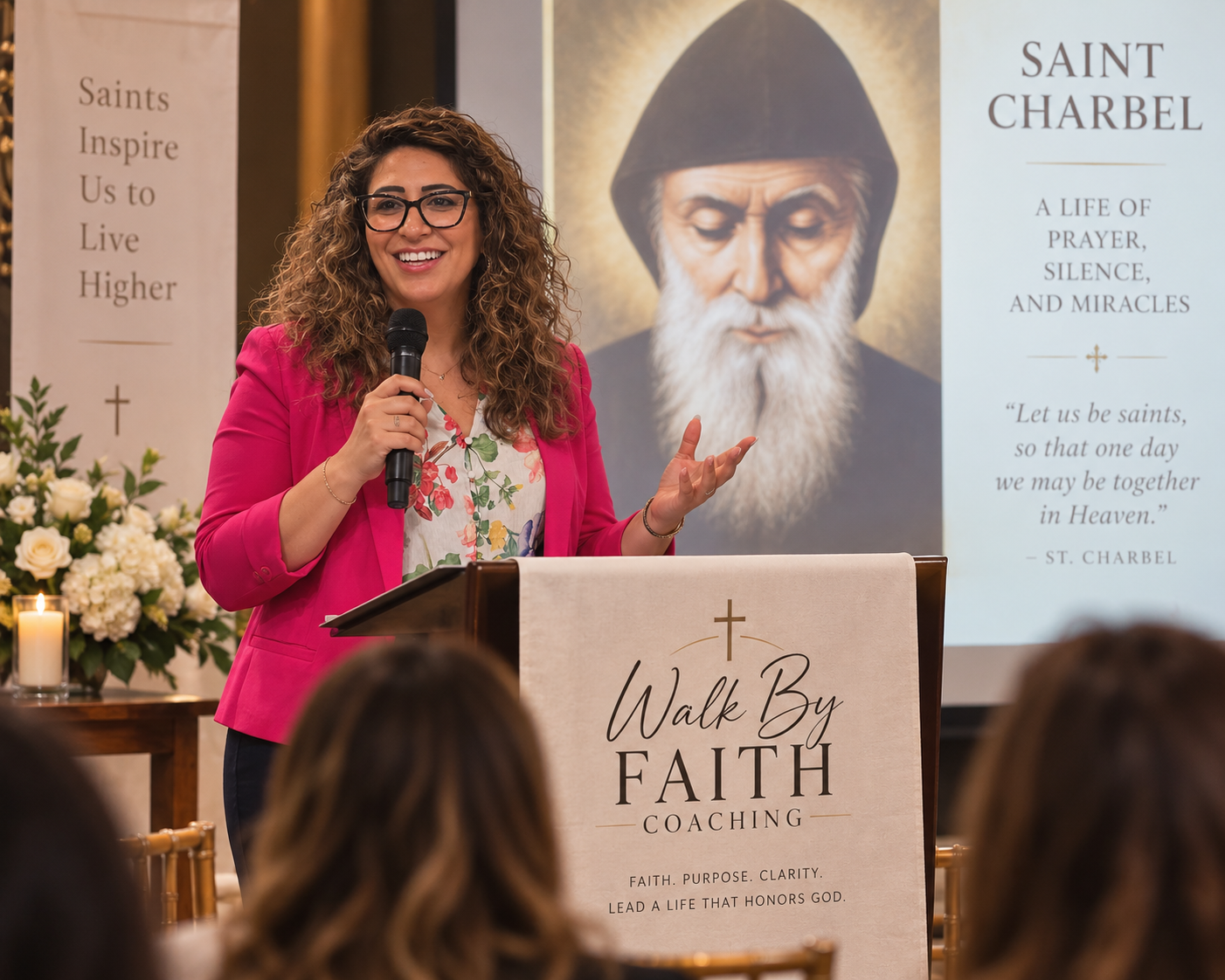A woman in glasses and a pink blazer is speaking into a microphone at a faith coaching event, standing behind a podium with a banner that reads "Walk By Faith Coaching." In the background, a large poster features an image of Saint Charbel and quotes about his life of prayer and miracles.