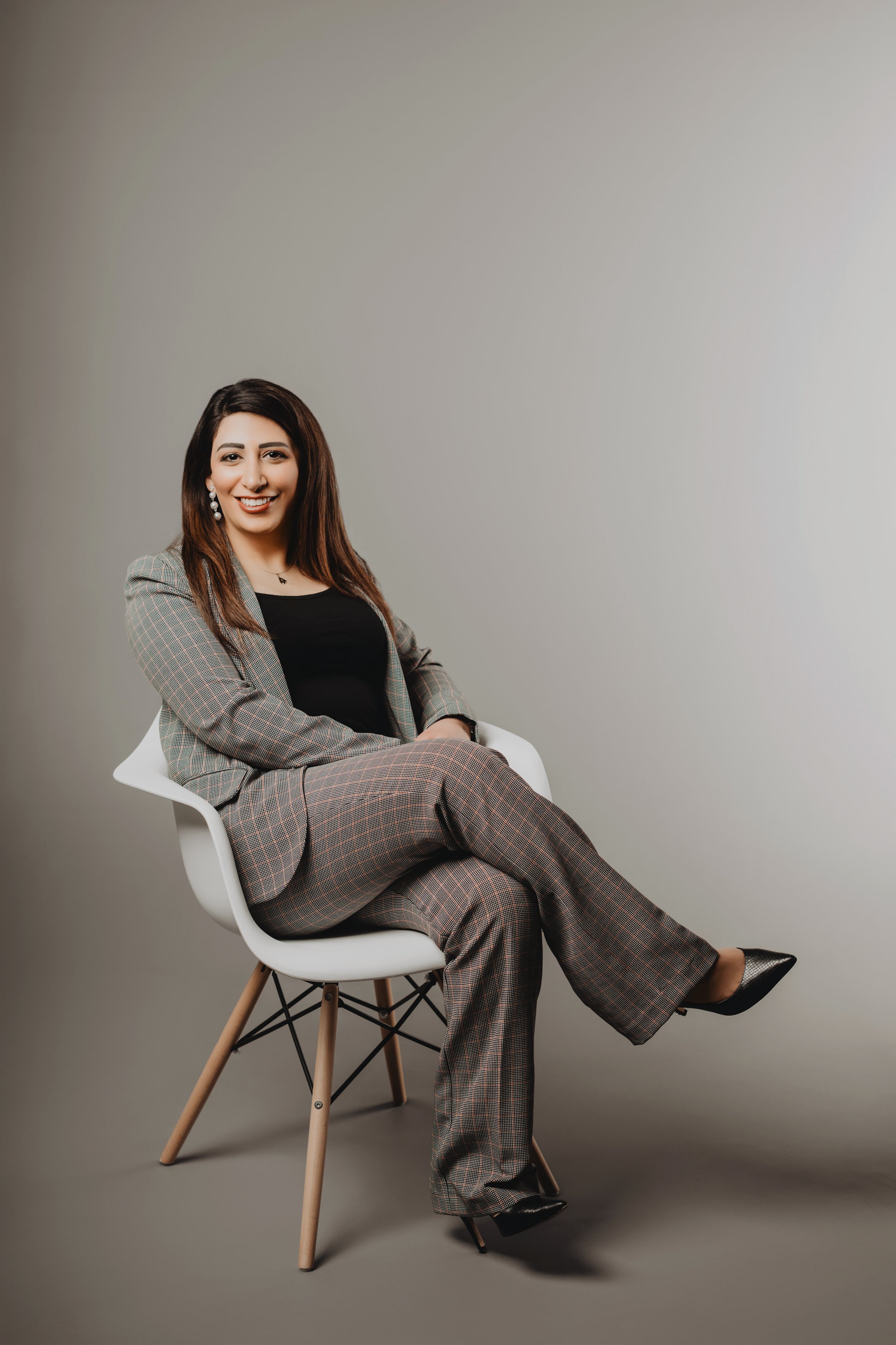 A woman with long dark hair wearing a plaid suit, black top, and black heels, sitting in a white chair against a plain gray background, smiling and looking at the camera.