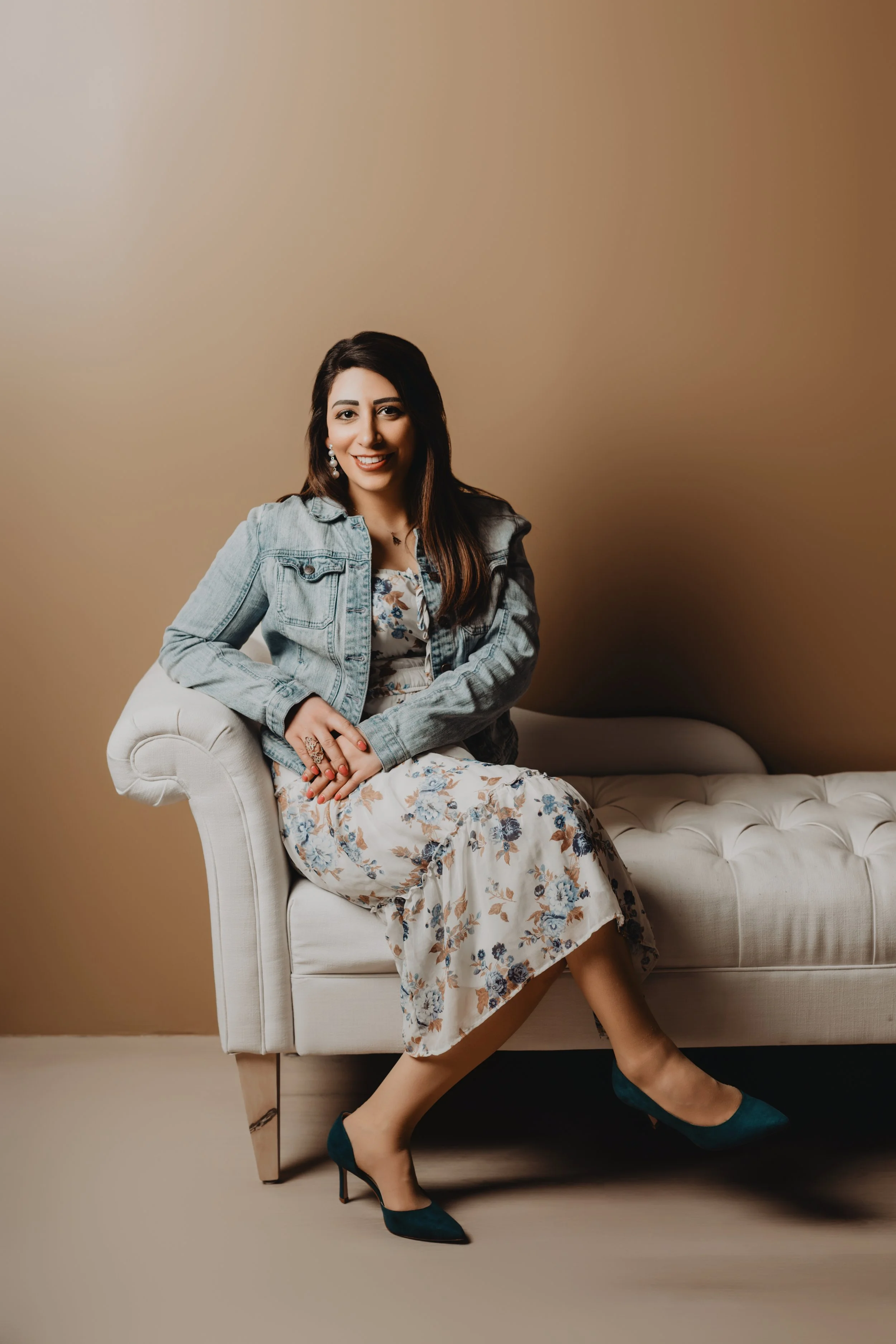 A woman with long dark hair, wearing a denim jacket, floral dress, and teal high heels, sitting sideways on a white tufted sofa against a plain brown wall, smiling and looking at the camera.