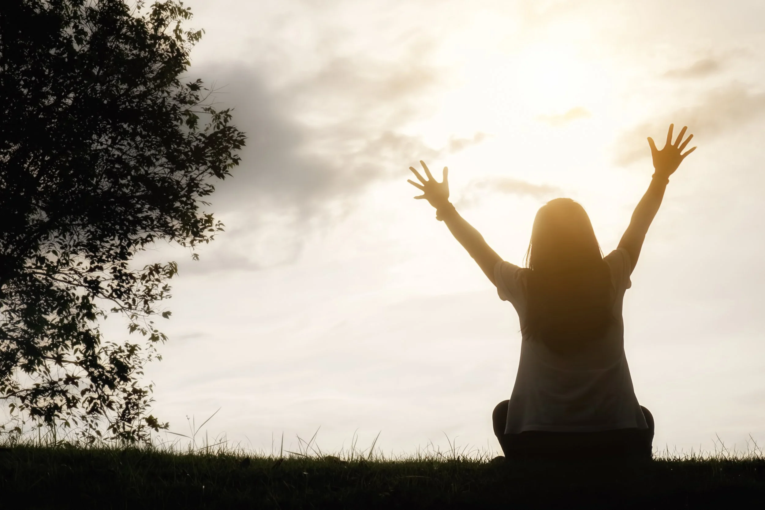 A person is sitting outdoors at sunset with their arms raised towards the sky, silhouetted against a cloudy sky with the sun shining through, near a tree.