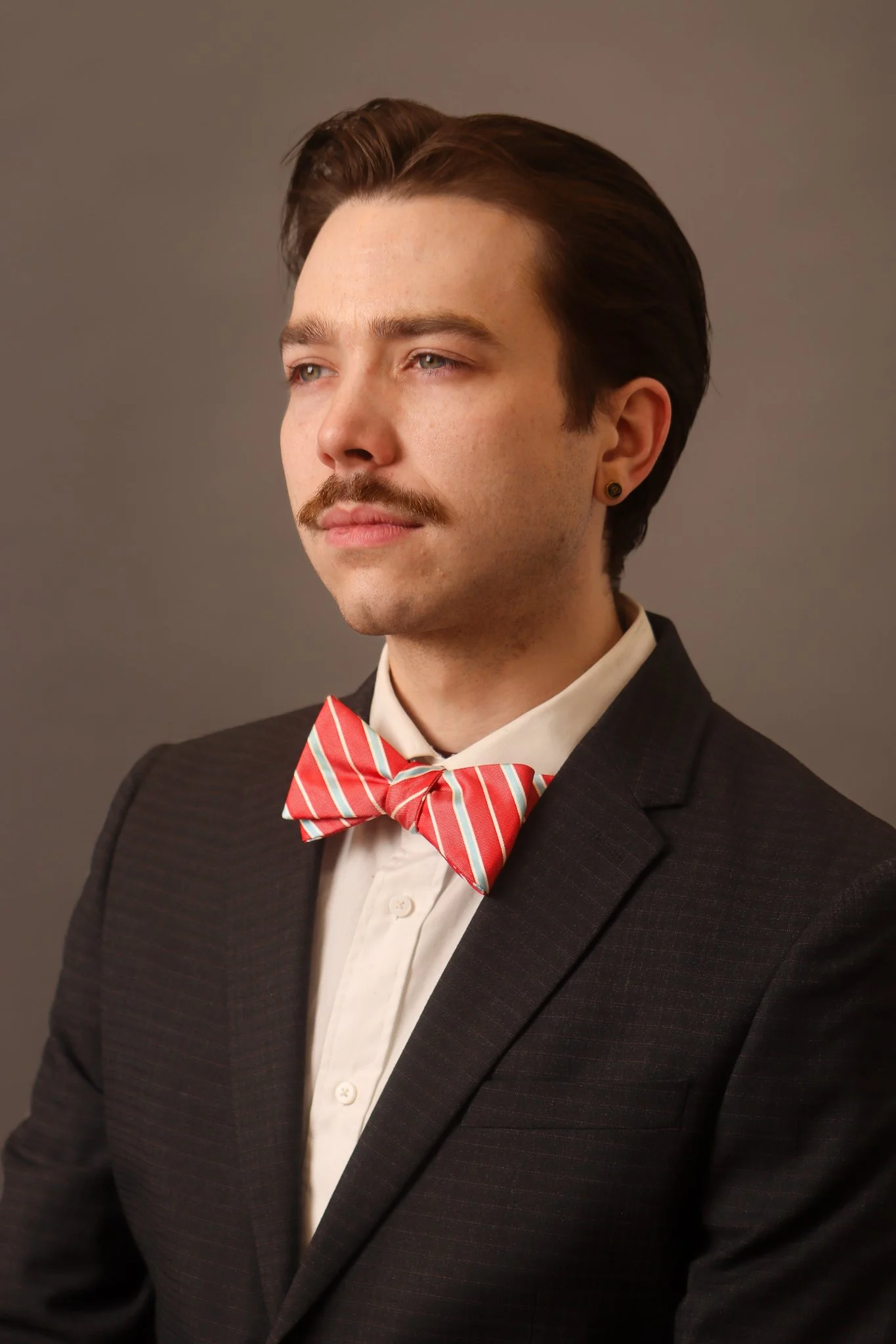 A man with brown hair and a mustache wearing a black suit, white shirt, and red striped bow tie, looking to the side against a plain background.
