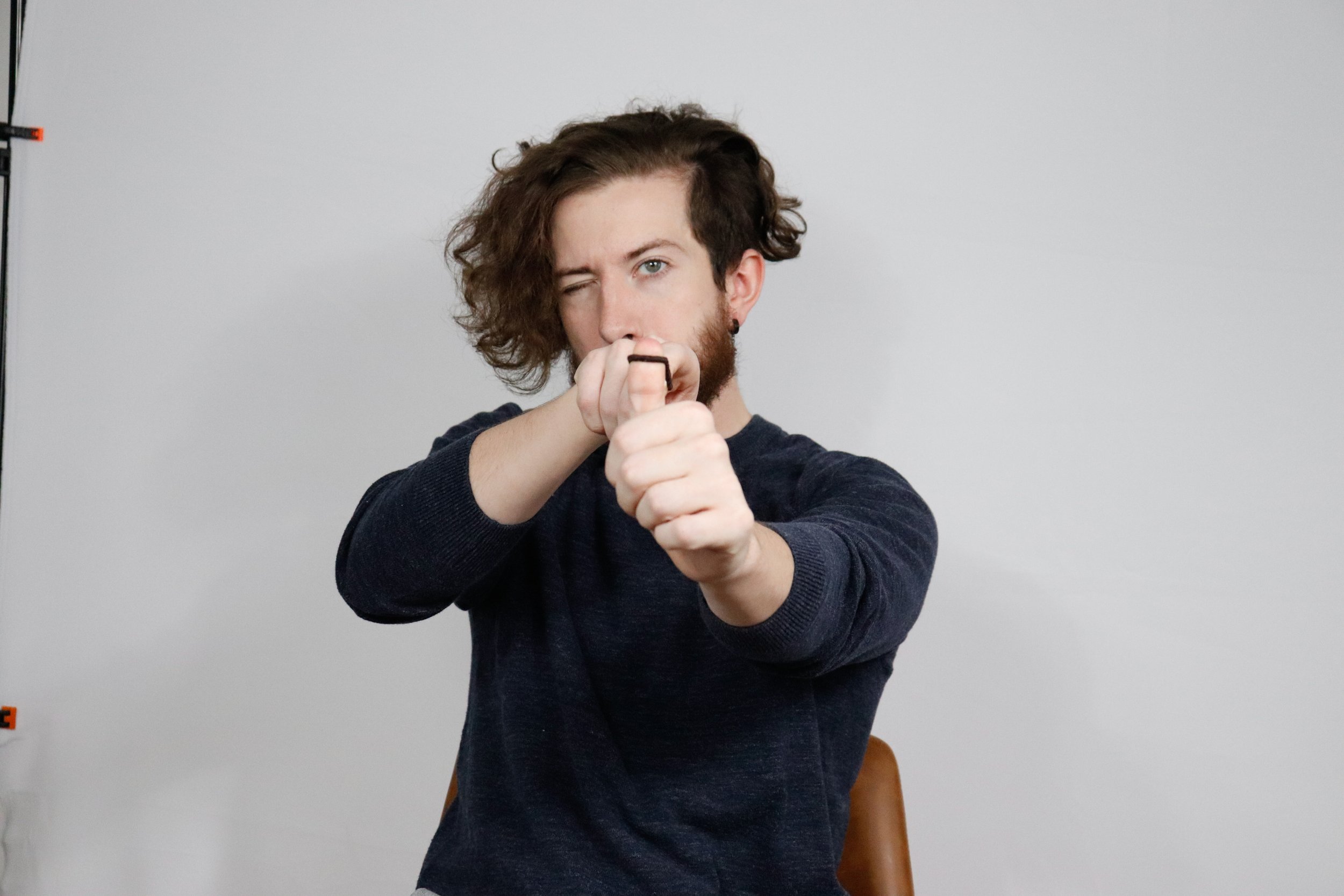 A young man with wavy brown hair and a beard is pointing directly at the camera with both hands, one above the other. He is wearing a dark long-sleeved shirt and standing against a plain light gray background.