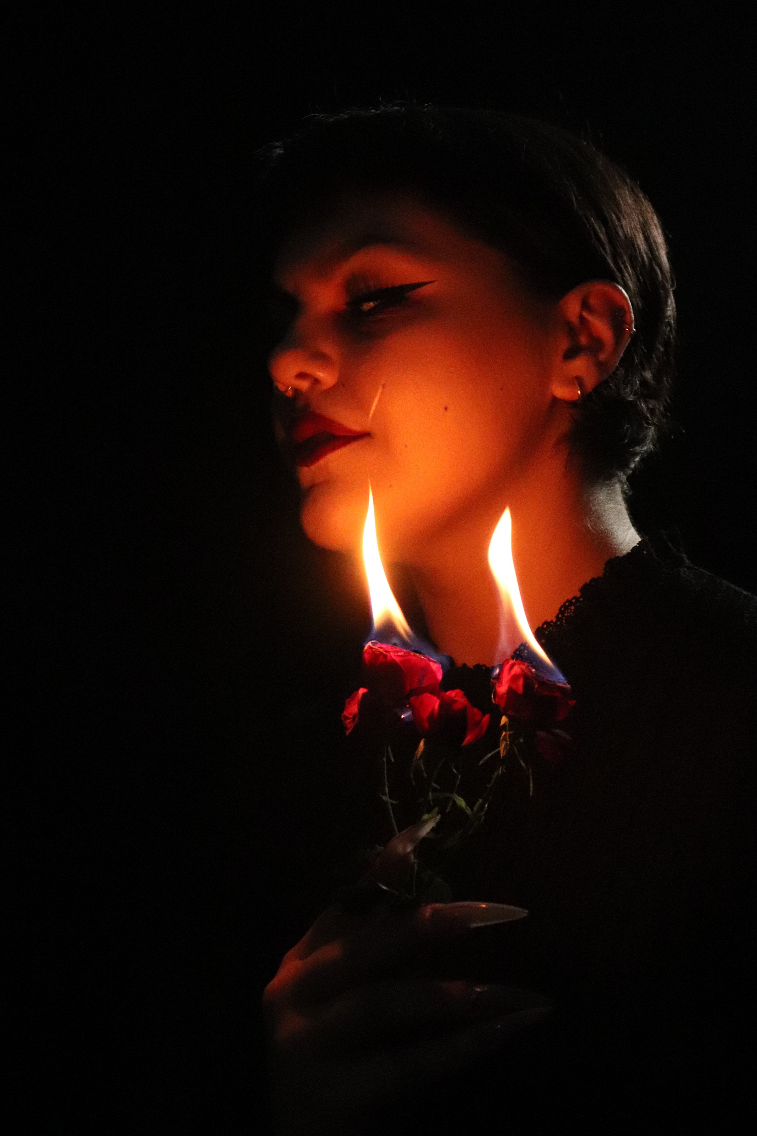 A person with short dark hair and multiple earrings holds a bouquet of red roses, with their face partially illuminated by the flames of burning roses, against a dark background.