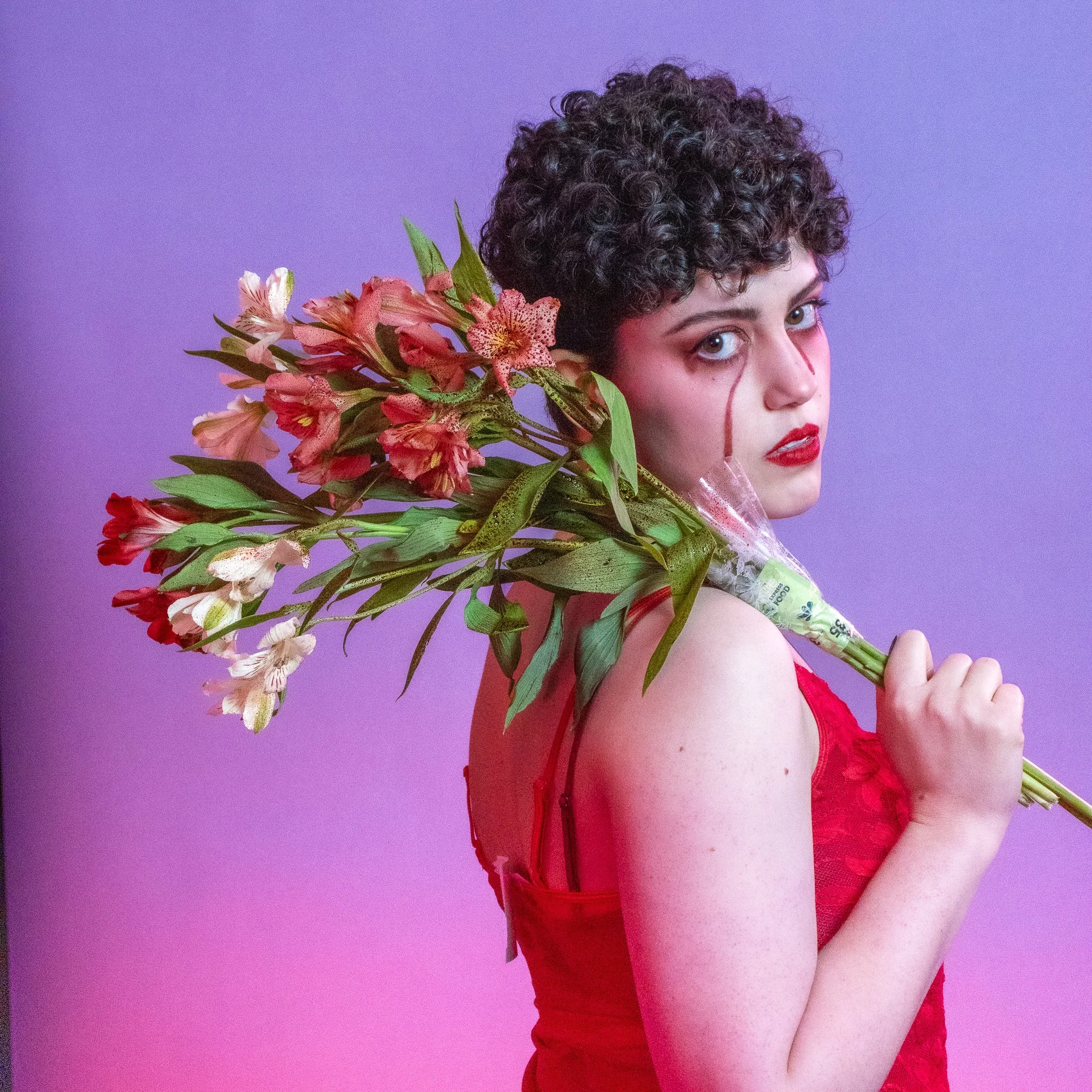 A woman with short curly dark hair wearing a red lace dress, holding a bouquet of pink and white flowers, against a purple background.