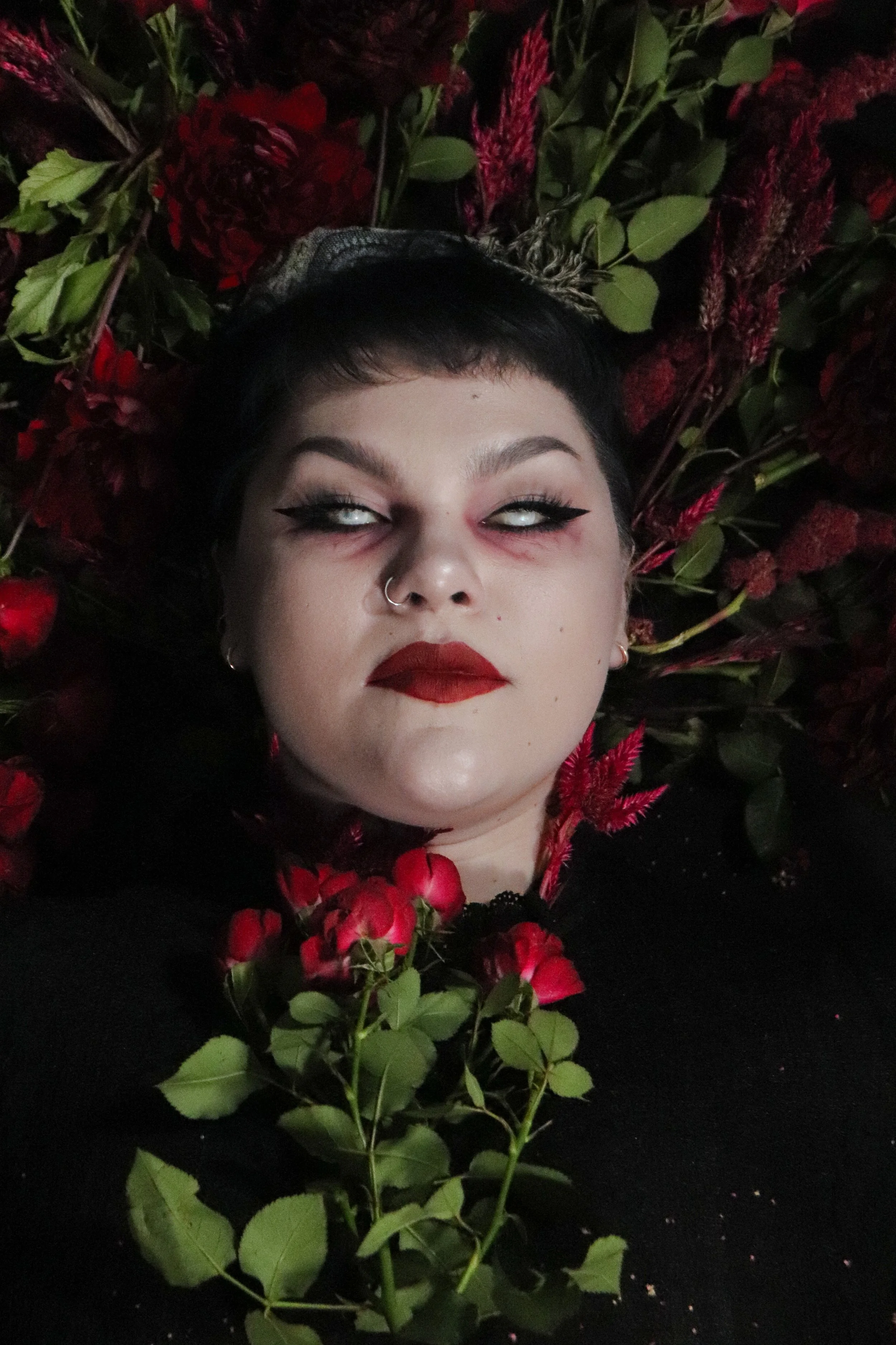 Person with dark hair and makeup lying among red roses and green leaves, looking up with eyes rolled back, dark eyeliner, red lipstick, and a septum piercing.