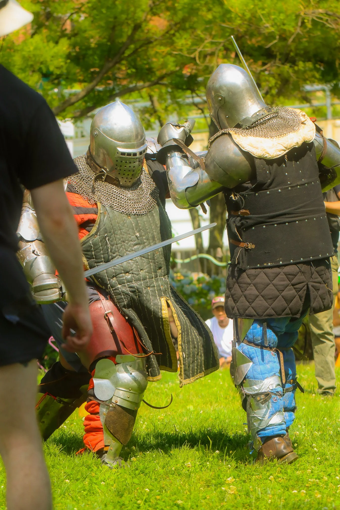 Two individuals dressed as knights in medieval armor engaging in a reenactment or duel outdoors on a grassy field with trees in the background.