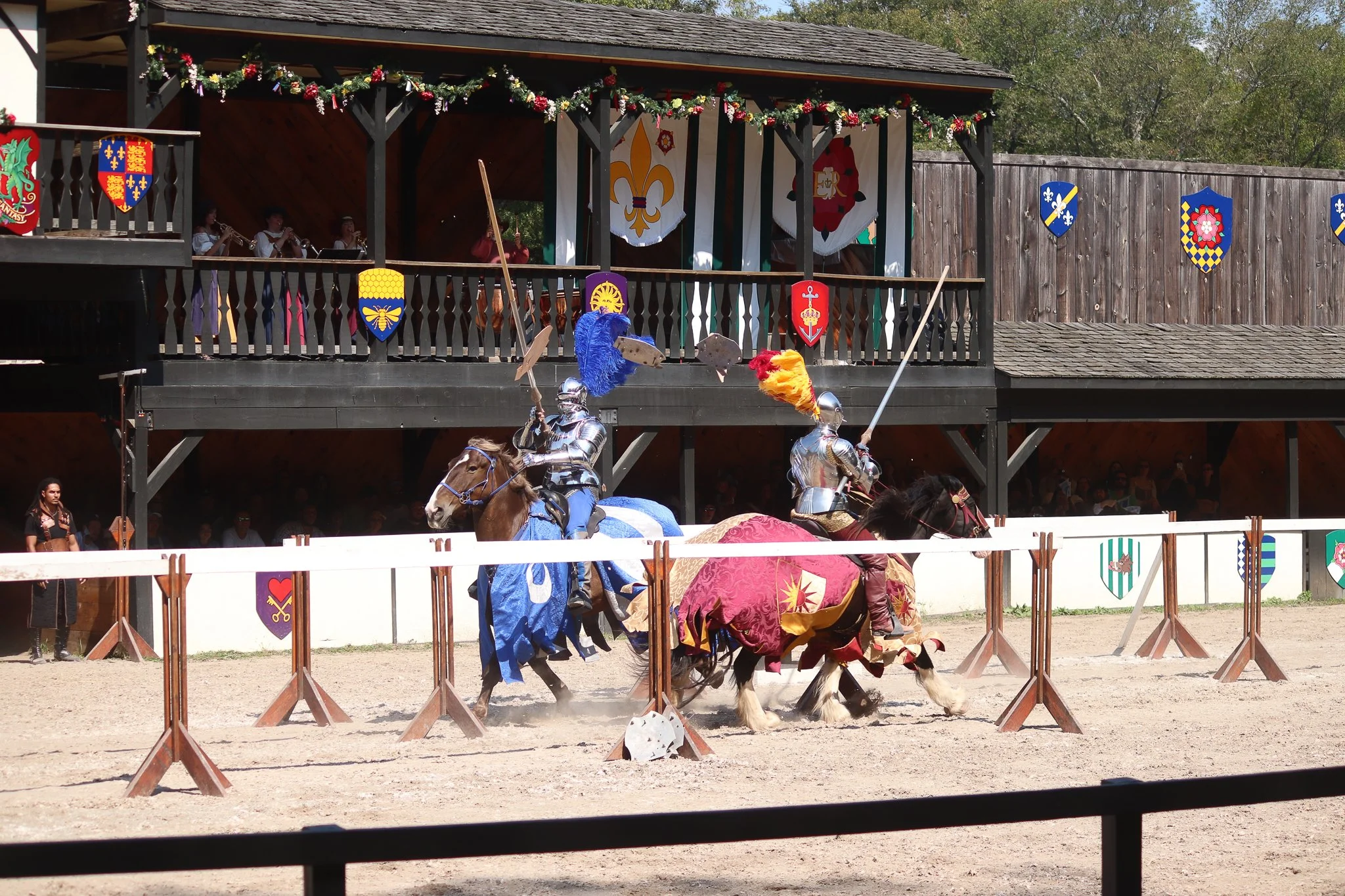 A medieval jousting tournament with armored knights on horses and raised shields, spectators in the background, and a second story with musicians and shield banners