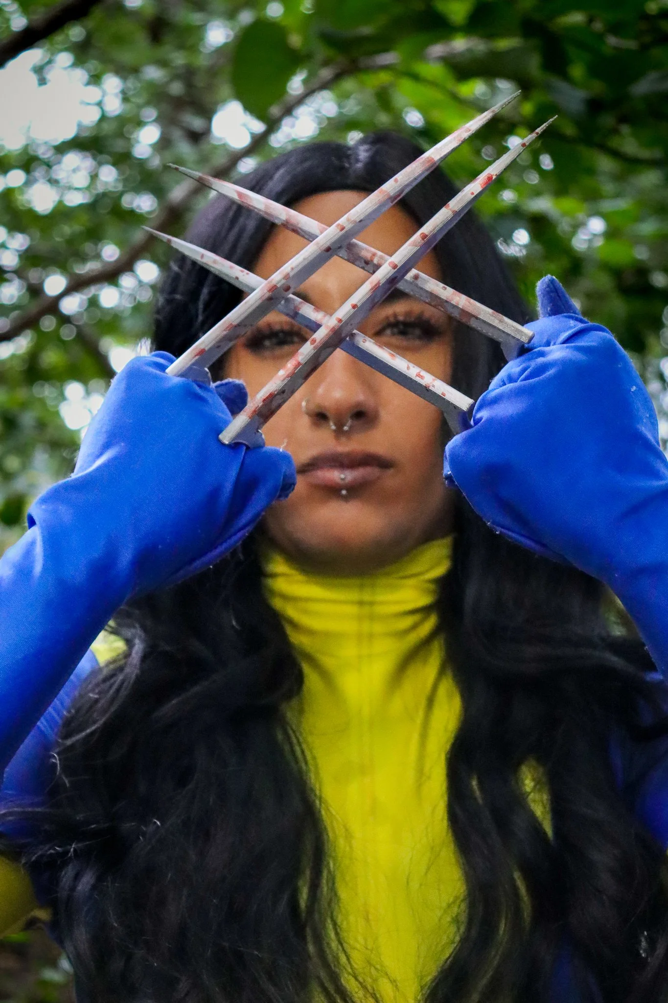 Person with long black hair, blue gloves, and yellow jacket holding four knives crossed in front of face, with a leafy green background.