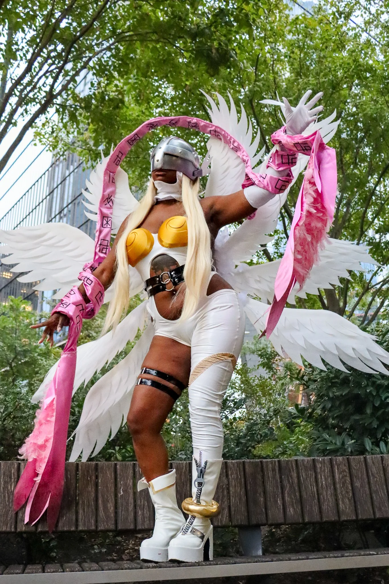 A performer dressed in a fantasy costume with large white wings, a silver helmet, pink ribbons, gold armor pieces, and futuristic white boots, standing outdoors in front of green trees and urban buildings.