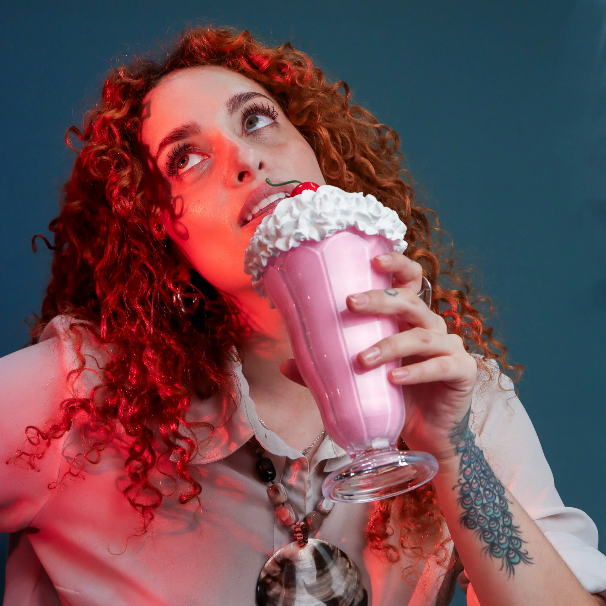 A woman with curly red hair holding a pink milkshake with whipped cream and a cherry on top, looking upwards.