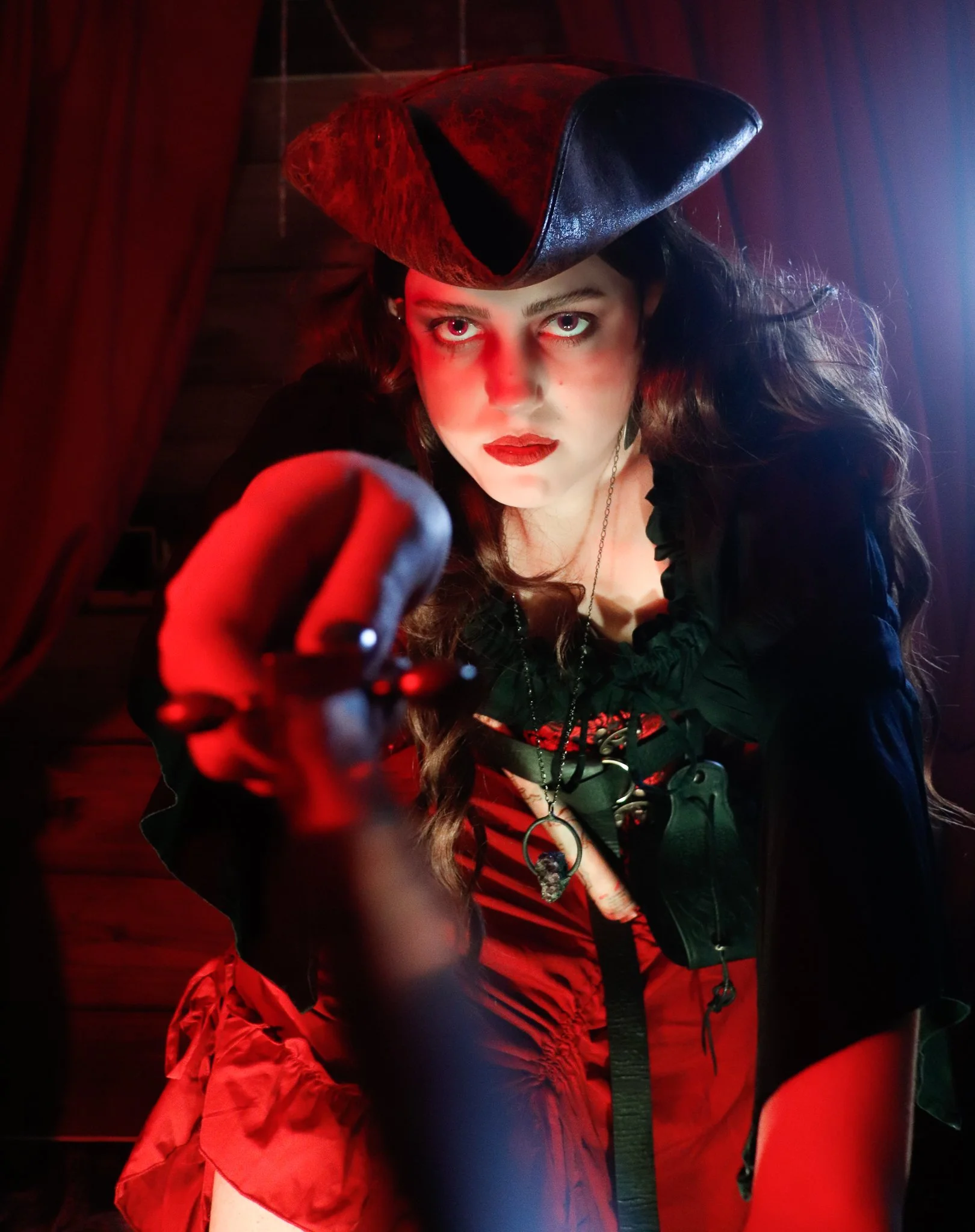 A woman with long dark hair and bright eyes looking directly at the camera, wearing a black hat with a wide brim, a necklace with a skull pendant, a black top, and red pants. The photo is taken from above, with red lighting highlighting her face and 