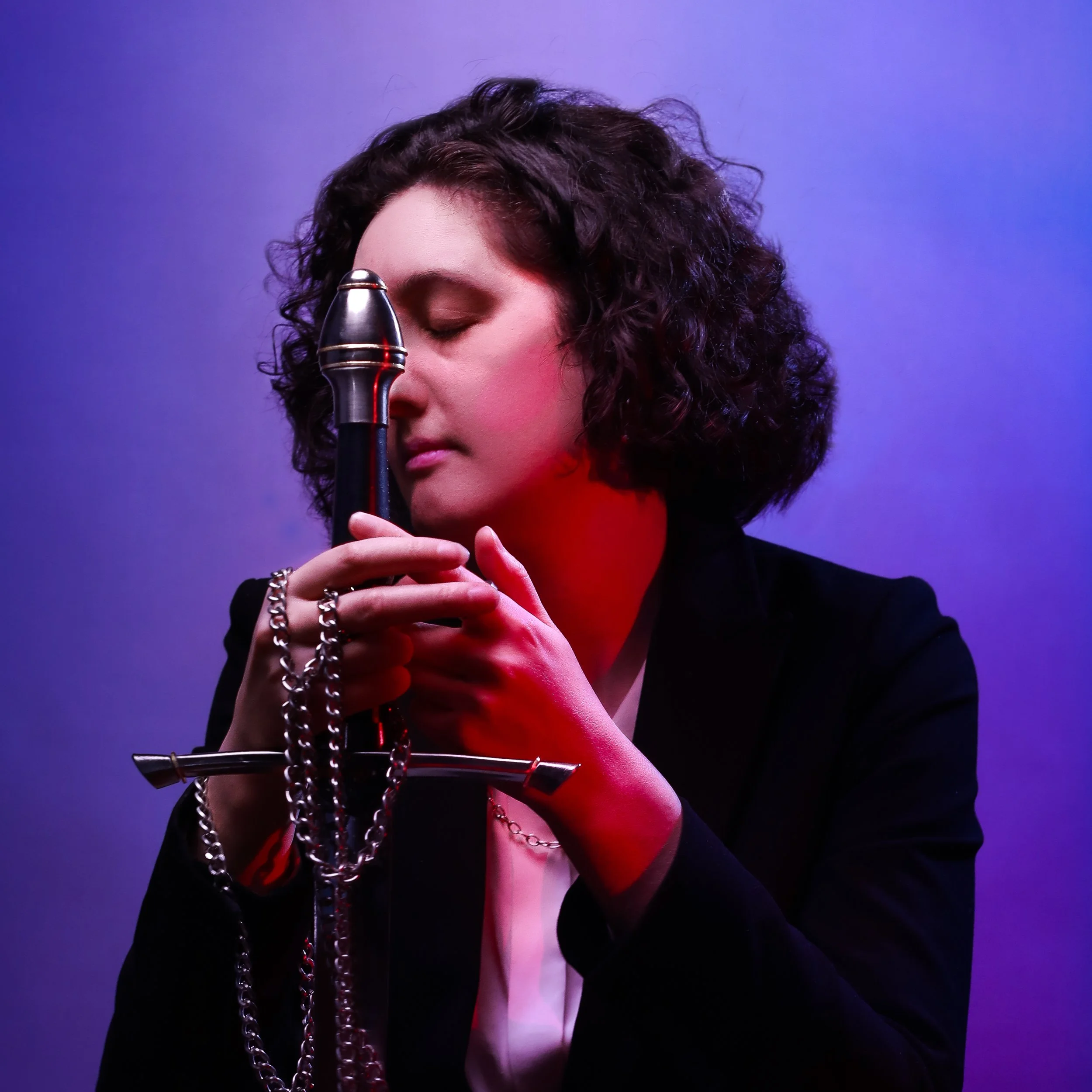 A woman with curly dark hair and makeup, holding a microphone close to her face with her eyes closed, against a colorful purple and blue gradient background.