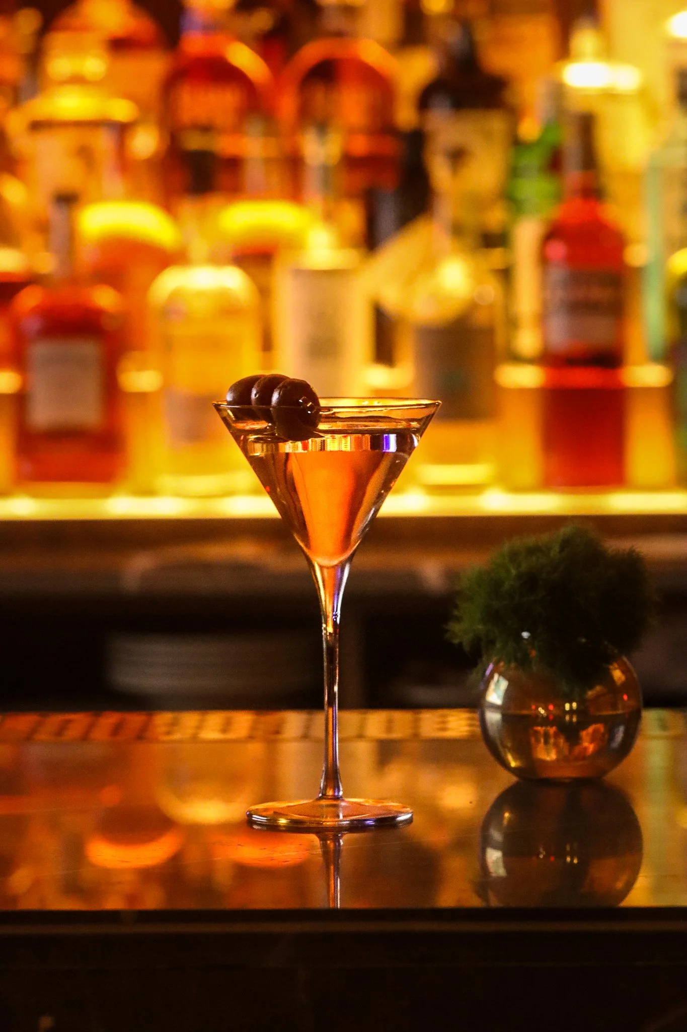 A martini glass with an olive garnish on top, set against a blurred background of colorful bottles lined on a bar shelf.
