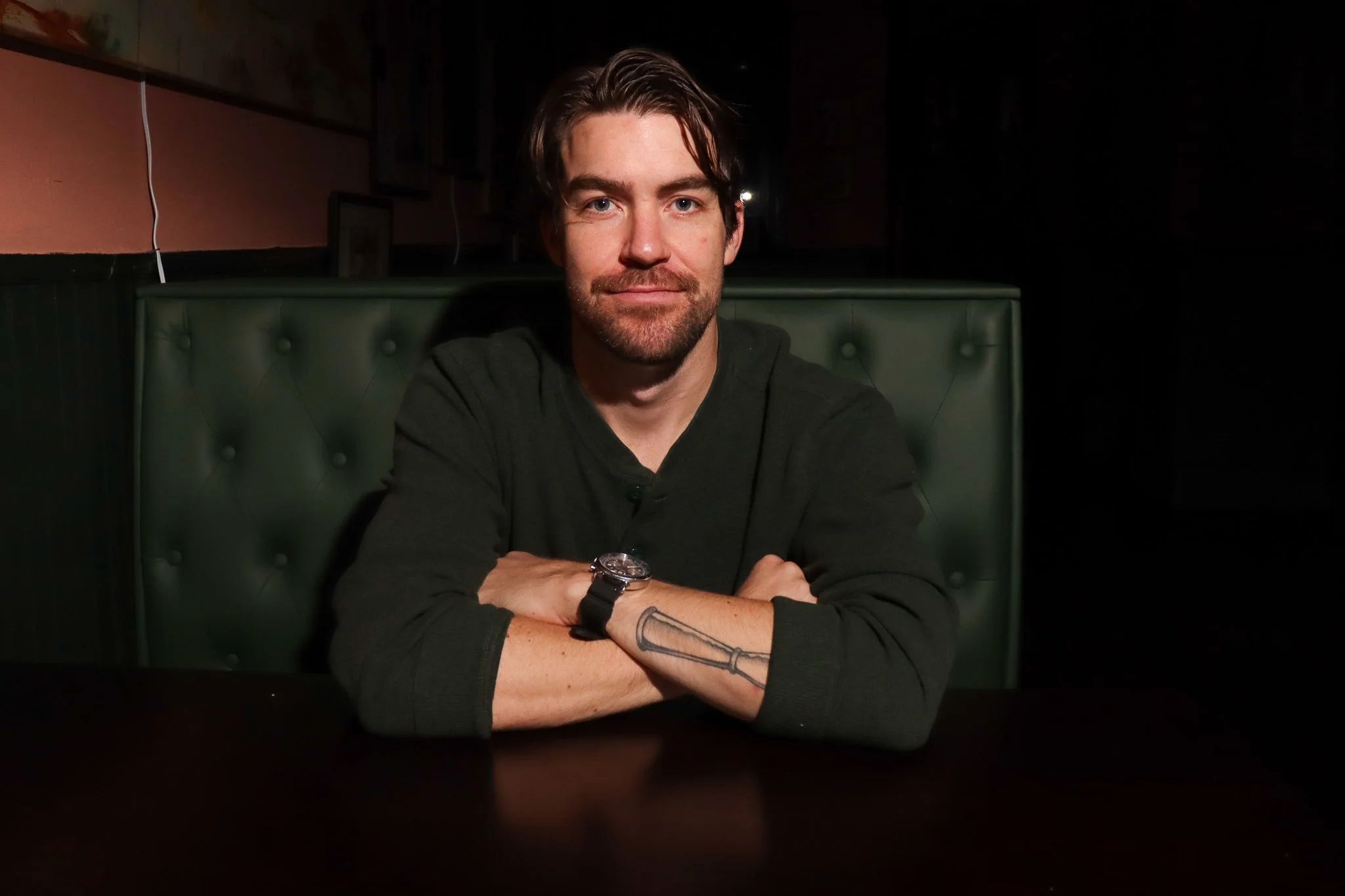 Man with brown hair, beard, and tattoos on his left arm sitting at a dark table in a dimly lit setting with green booth seating.